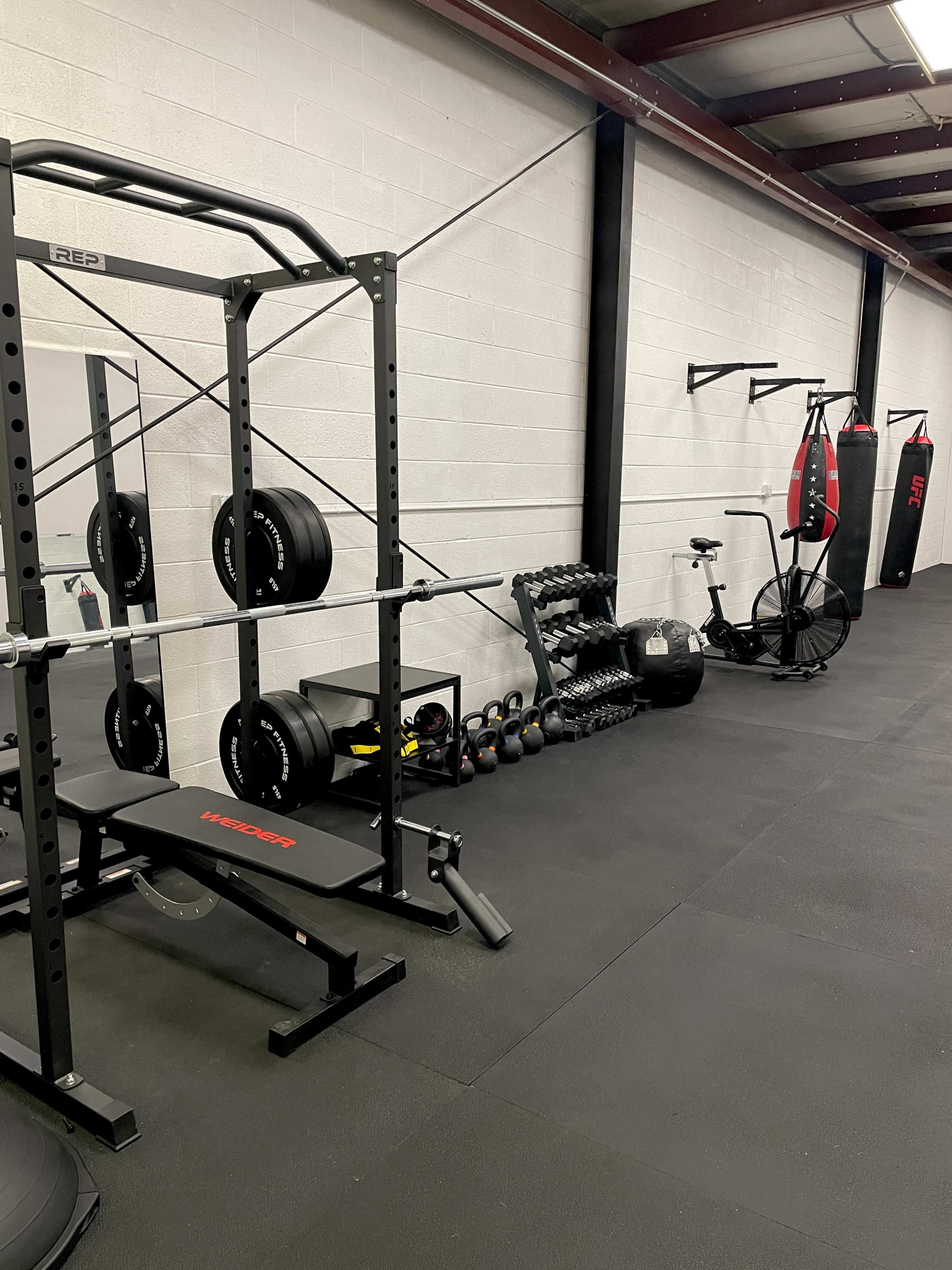 A gym interior featuring a squat rack with weights, a bench, a set of kettlebells, a water jug, a stationary bike, and a punching bag.