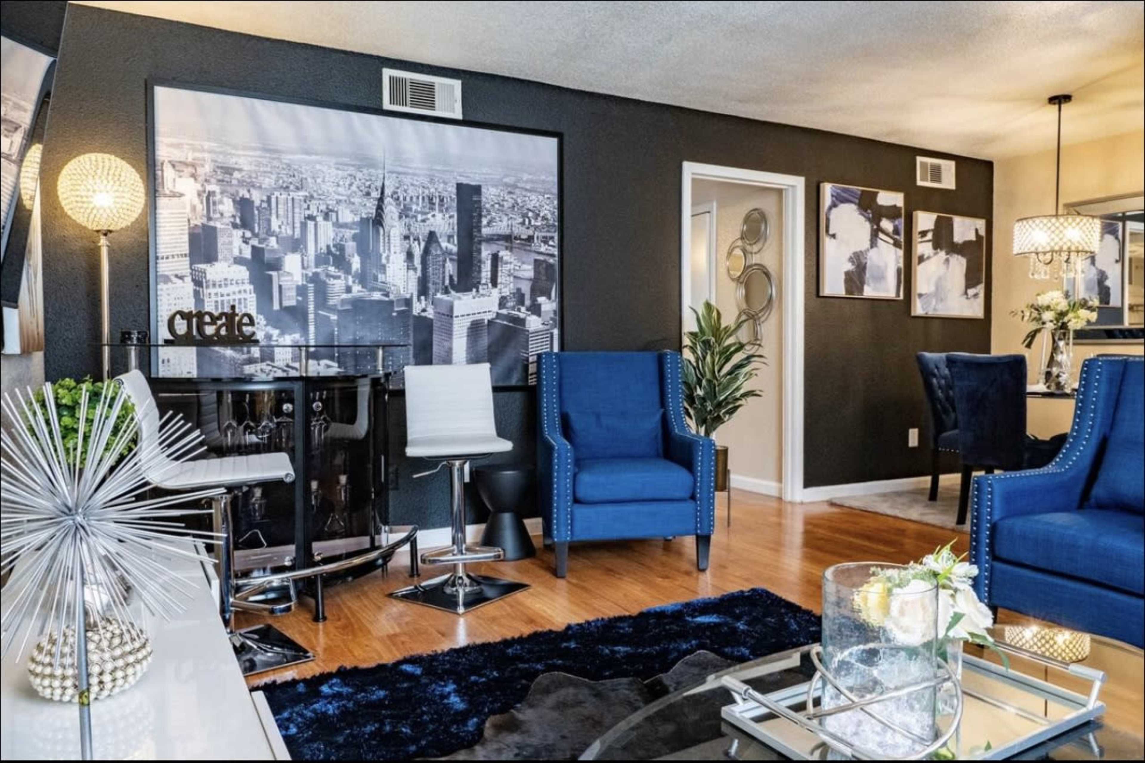 A modern living room features blue armchairs, a large cityscape wall art, and elegant decor elements, including a glass coffee table and stylish lighting.