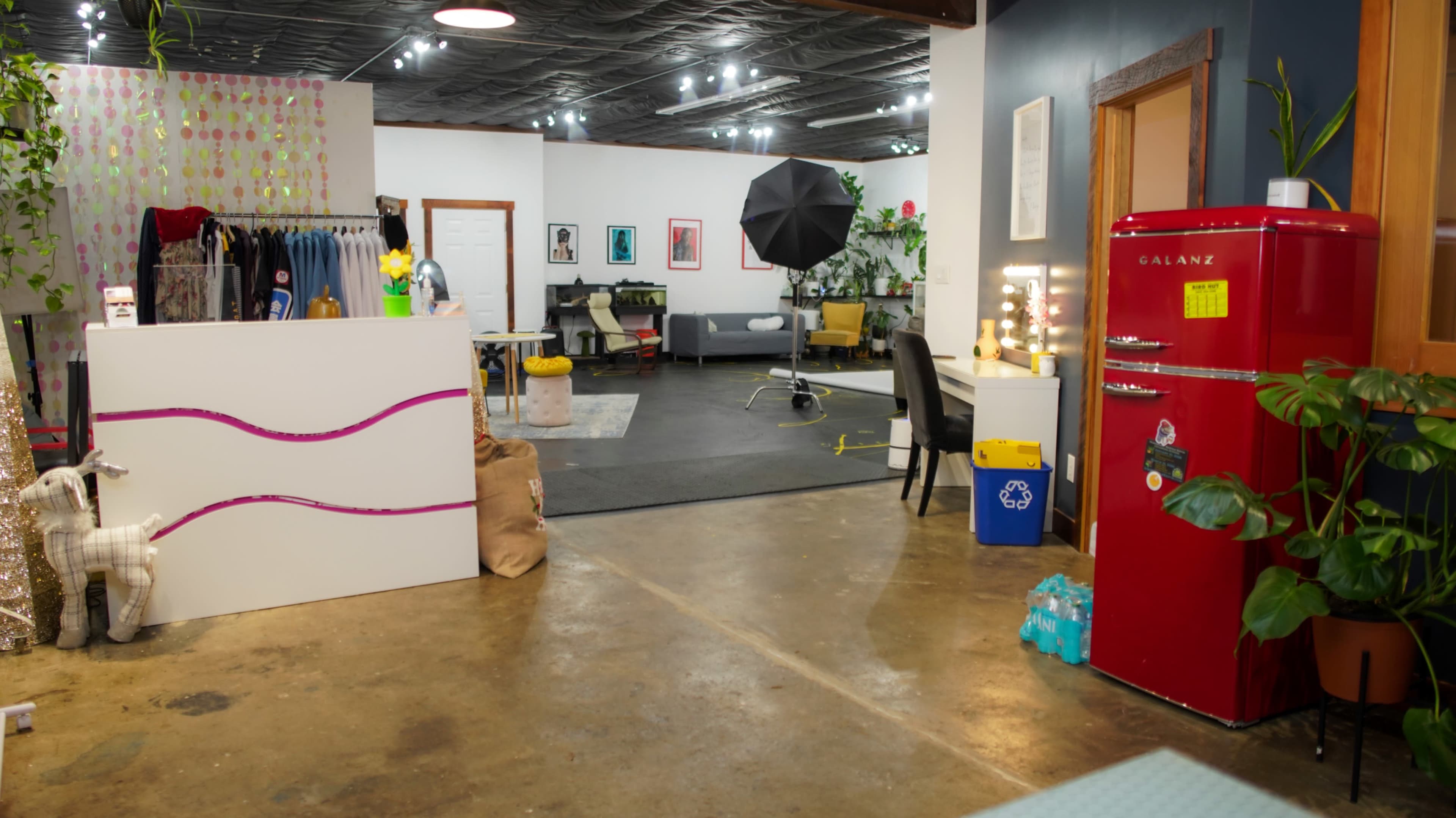 The scene features a spacious studio with a mix of clothing racks, a red refrigerator, a photography setup with an umbrella, and various furniture pieces arranged around a concrete floor.