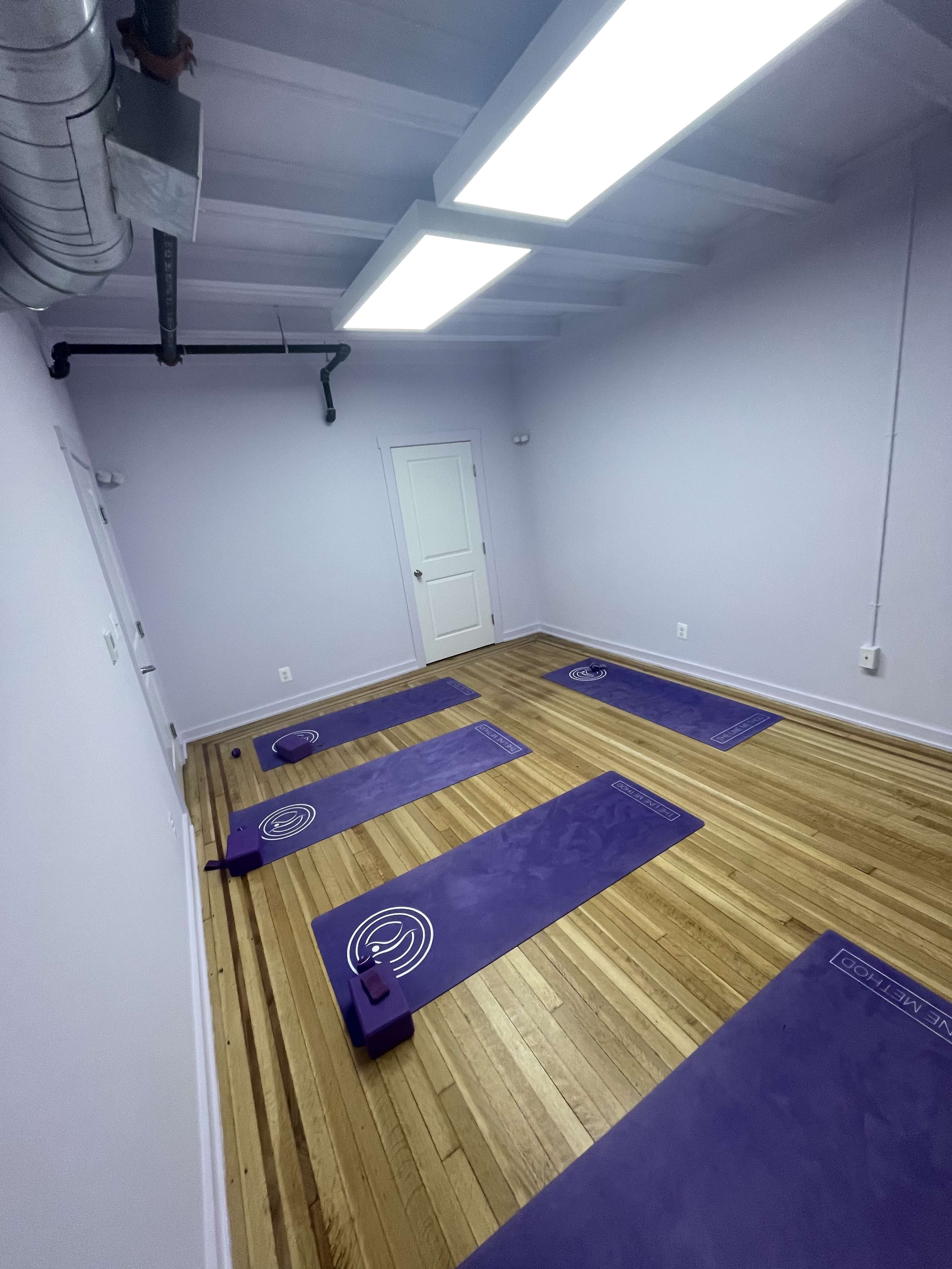 The image shows a yoga studio with purple mats and props arranged on a wooden floor, illuminated by overhead lights.