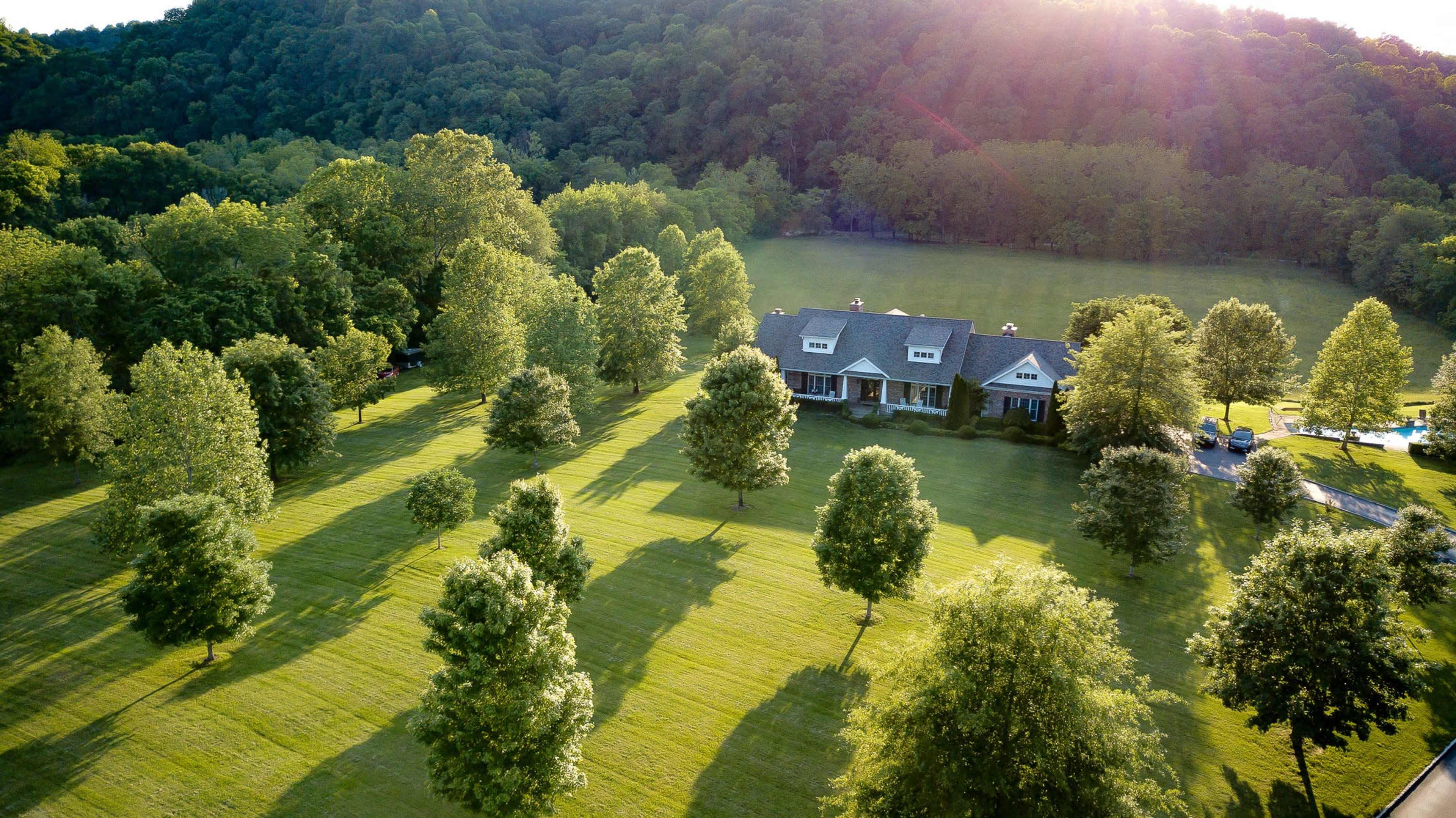 A large house is surrounded by a lush lawn and trees, set against a backdrop of rolling hills.