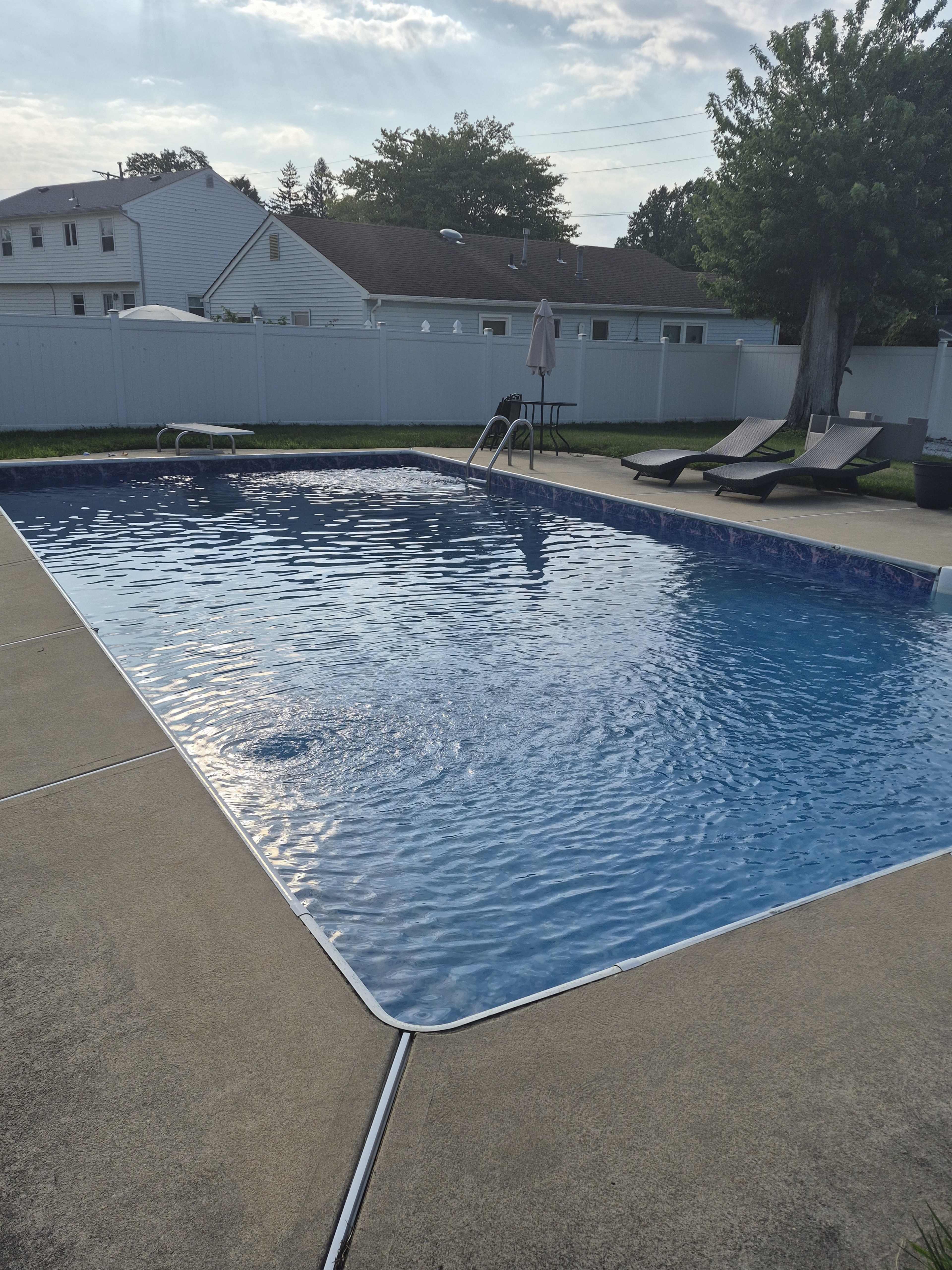 A clear blue swimming pool is surrounded by a concrete deck, lounge chairs, and a white fence.