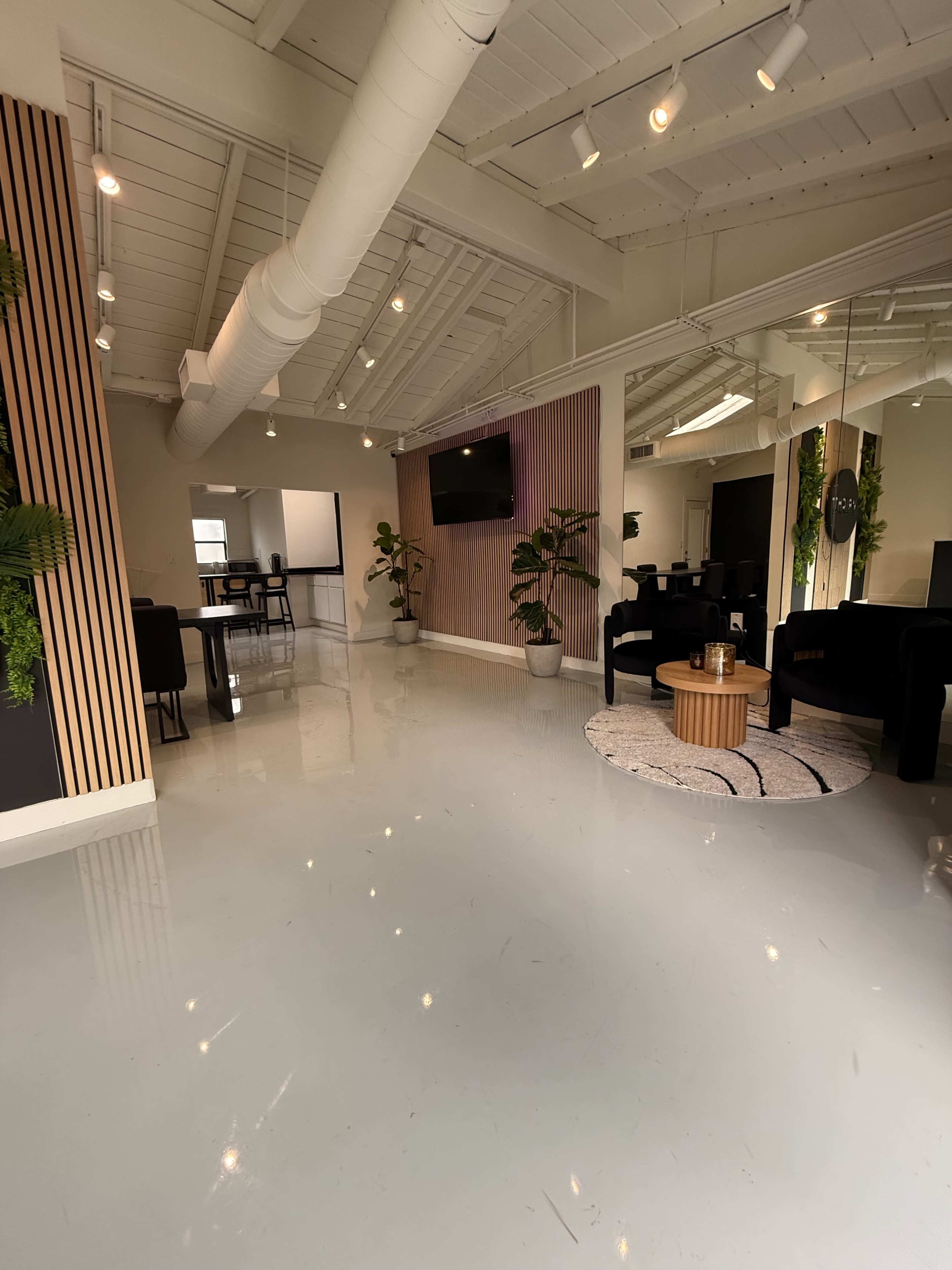 The image shows a modern, spacious office interior featuring a polished floor, seating areas, plants, and a wall-mounted television.