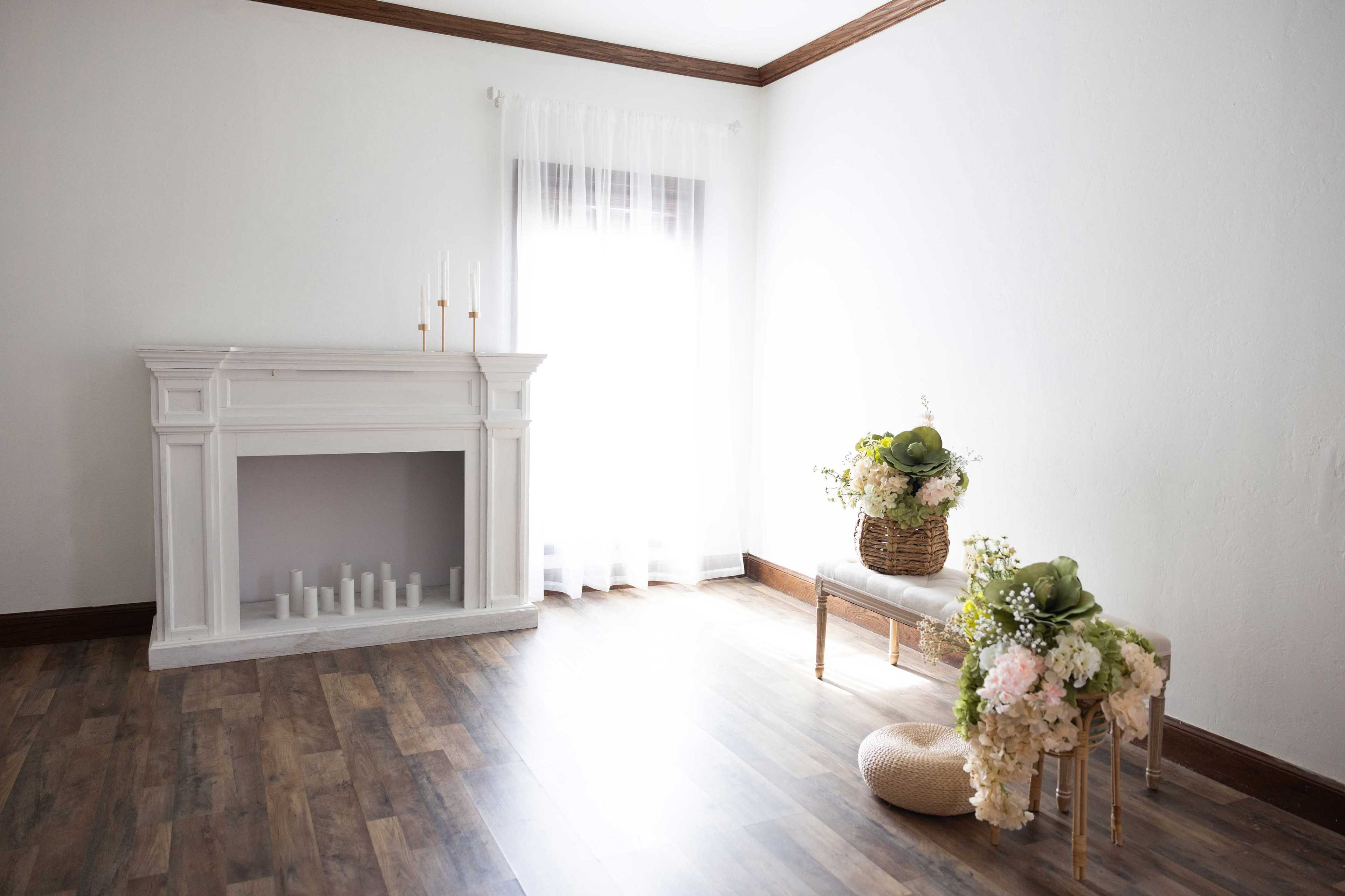 A bright, minimalist room features a white fireplace with candles and two decorative arrangements of flowers on a bench.