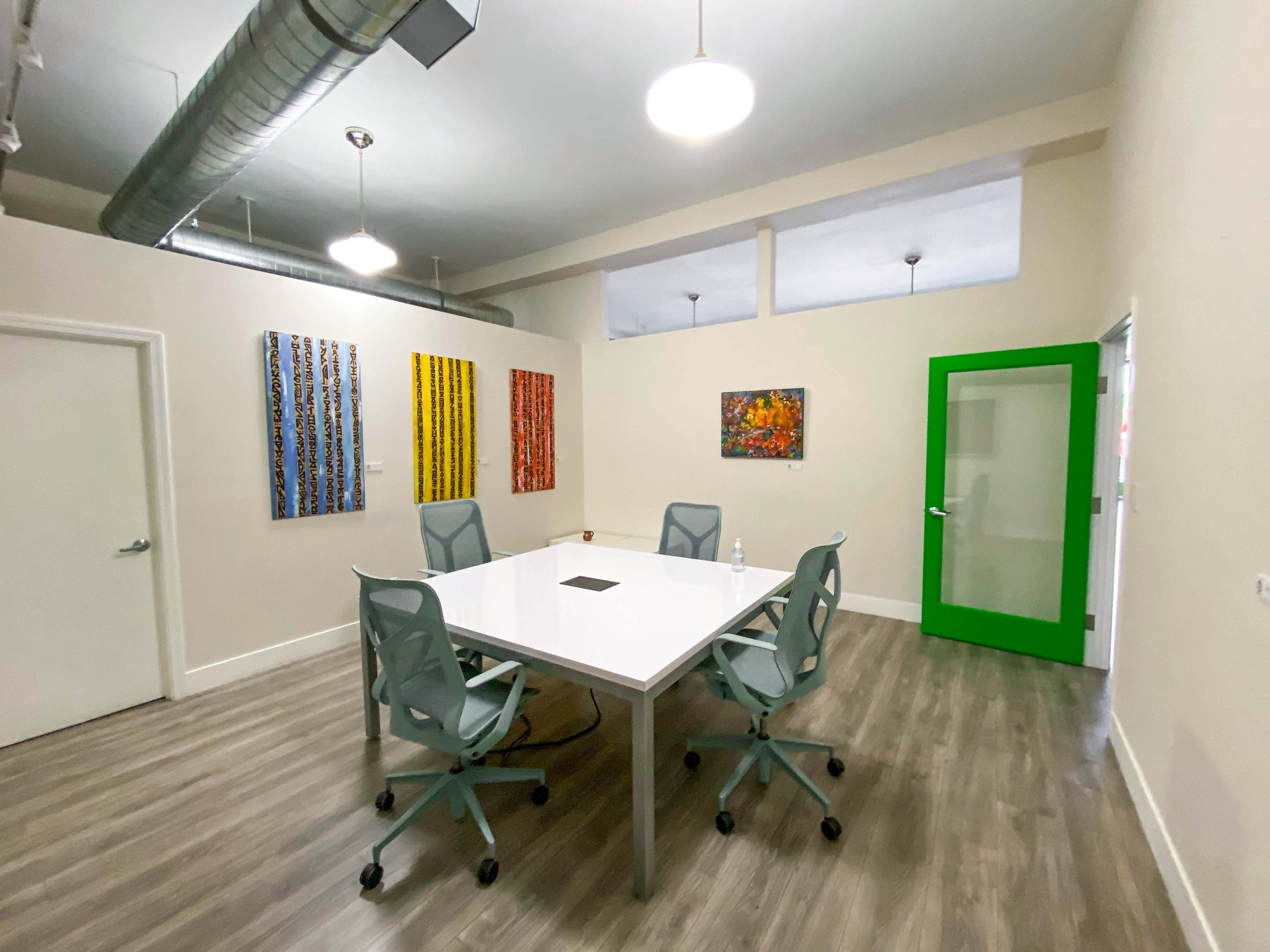 A modern conference room features a large white table surrounded by gray chairs, with colorful artwork displayed on the walls and a green glass door leading to another space.