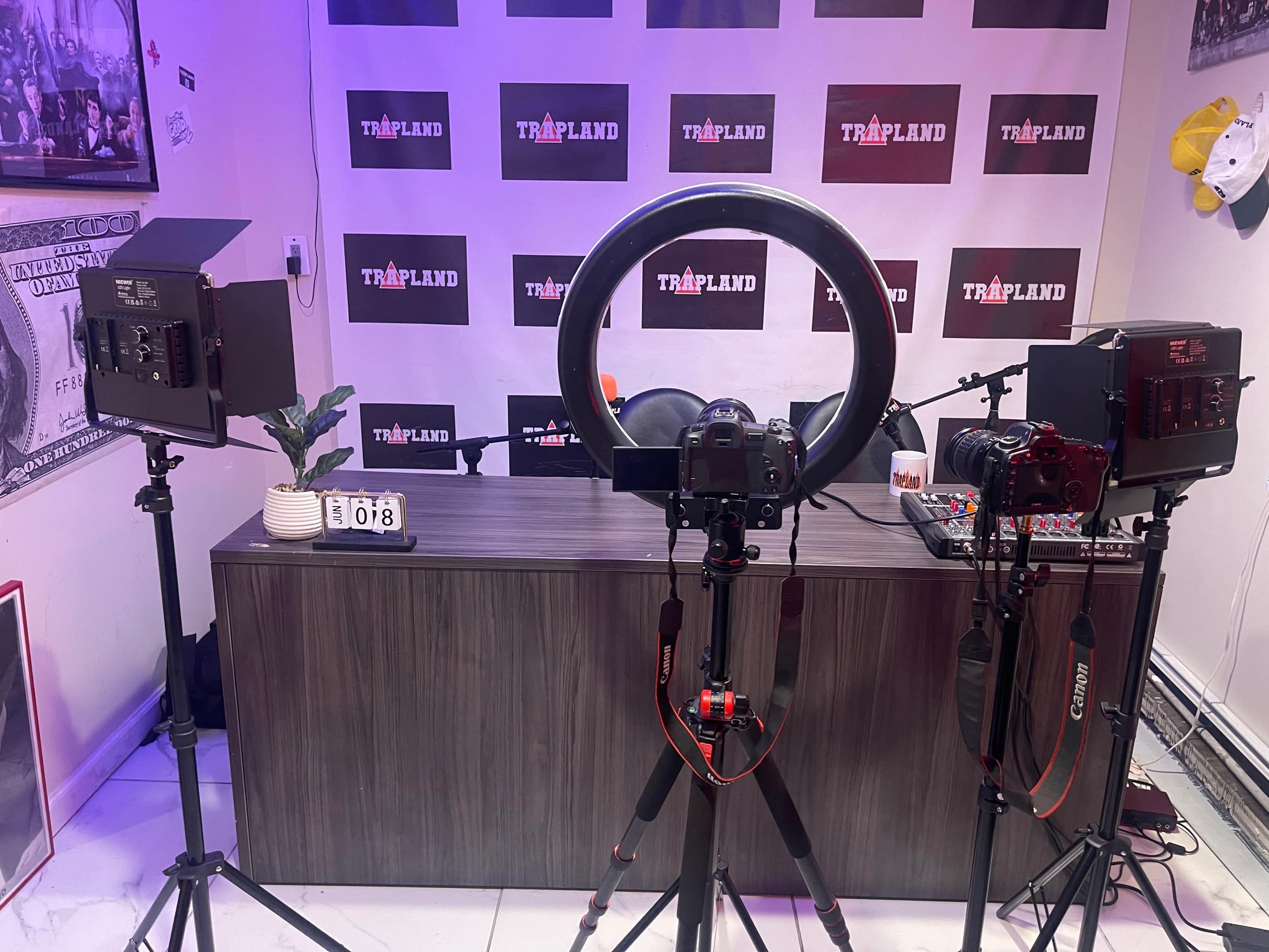 A video recording setup with two cameras, a ring light, a desk, and various lighting equipment arranged in front of a backdrop filled with the word "TRAPLAND."