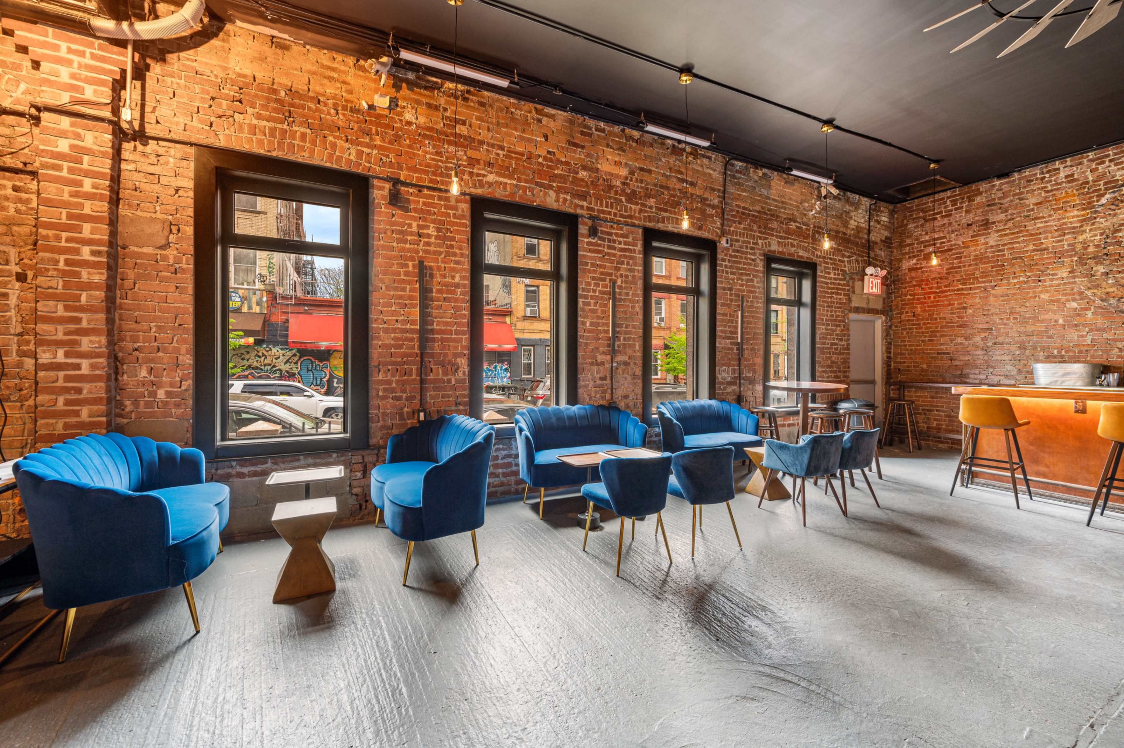A modern interior of a café or lounge featuring brick walls, blue upholstered chairs, wooden tables, and large windows.