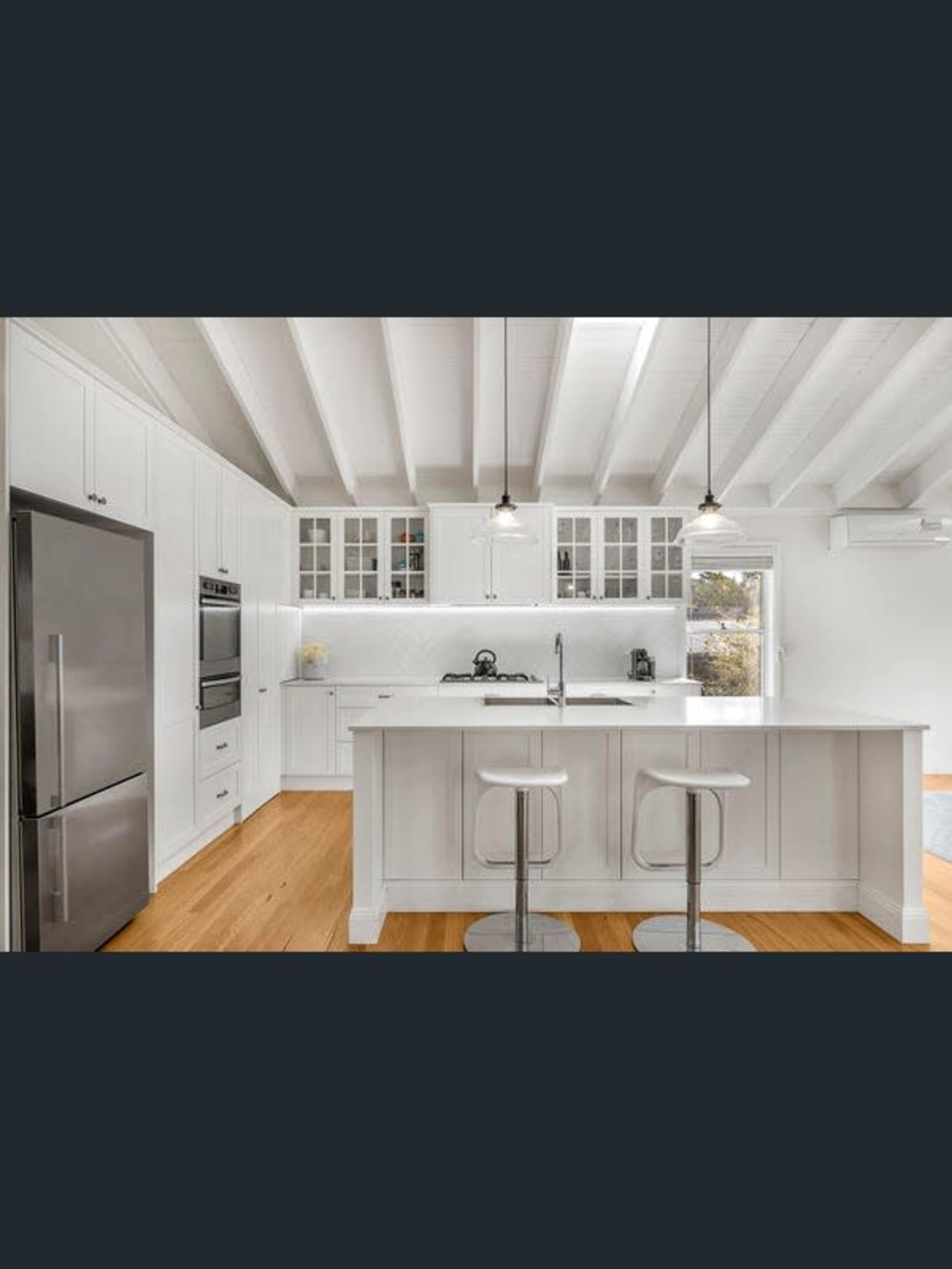 The image shows a modern kitchen with white cabinetry, stainless steel appliances, and a central island with two stools.