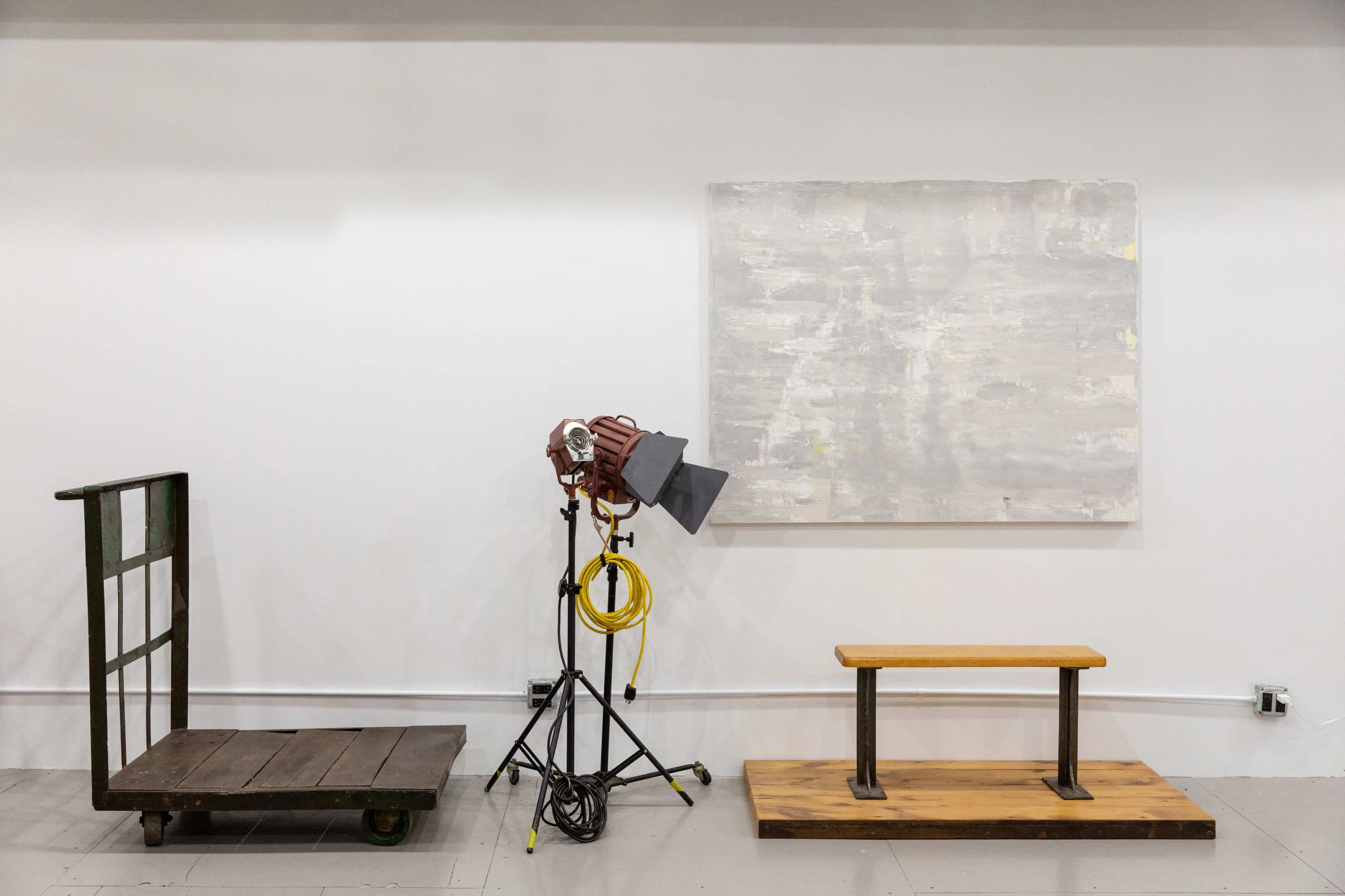 The image features an industrial cart and a wooden bench on a gray floor, accompanied by a lighting fixture on a tripod and a large abstract painting on the wall.