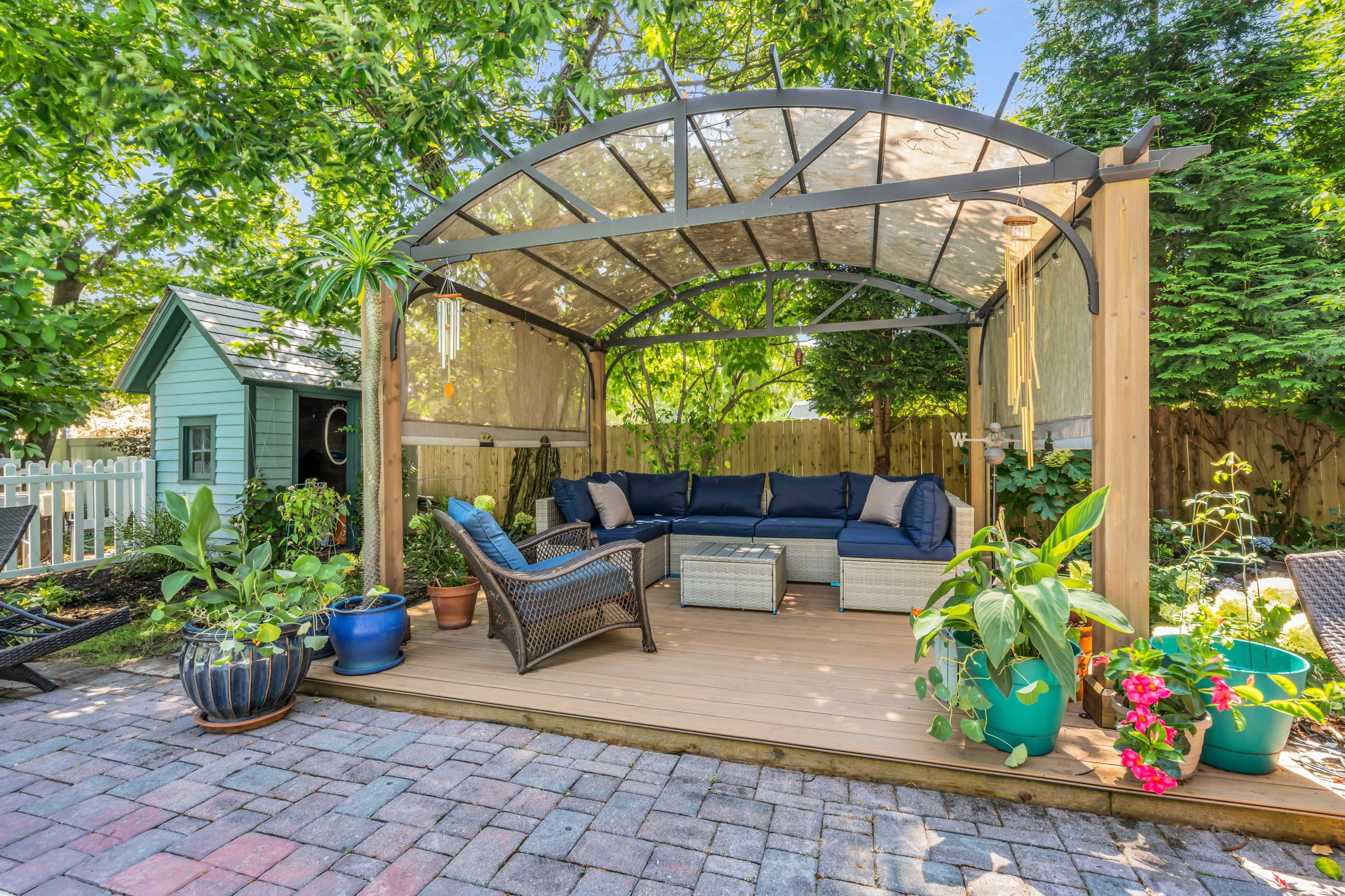 A covered outdoor seating area with a sectional sofa, coffee table, and various potted plants is surrounded by greenery and a small garden shed.