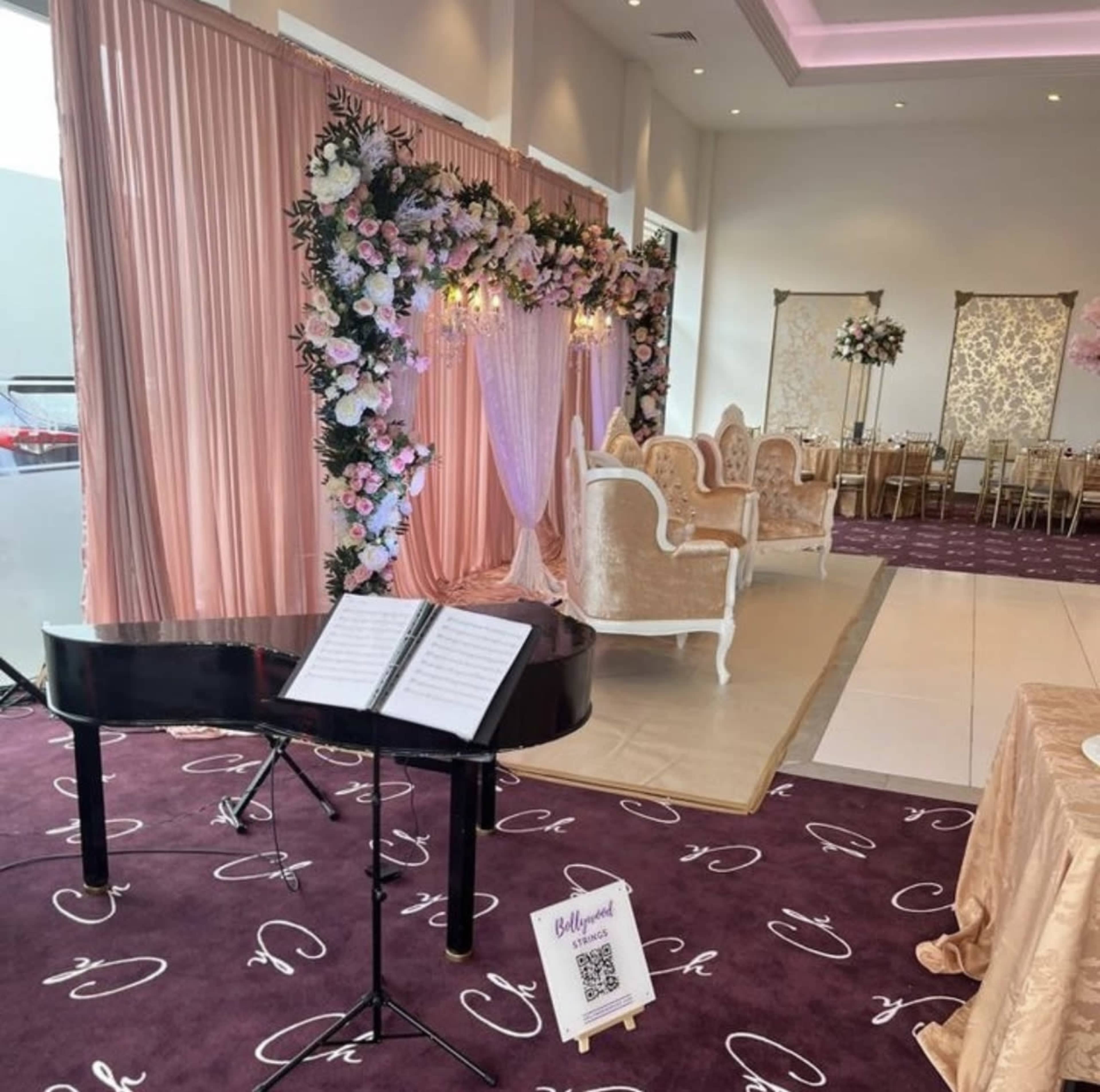 The image shows a decorated event space with a black piano and a backdrop of flowers, along with elegant seating arrangements in a light-colored setting.