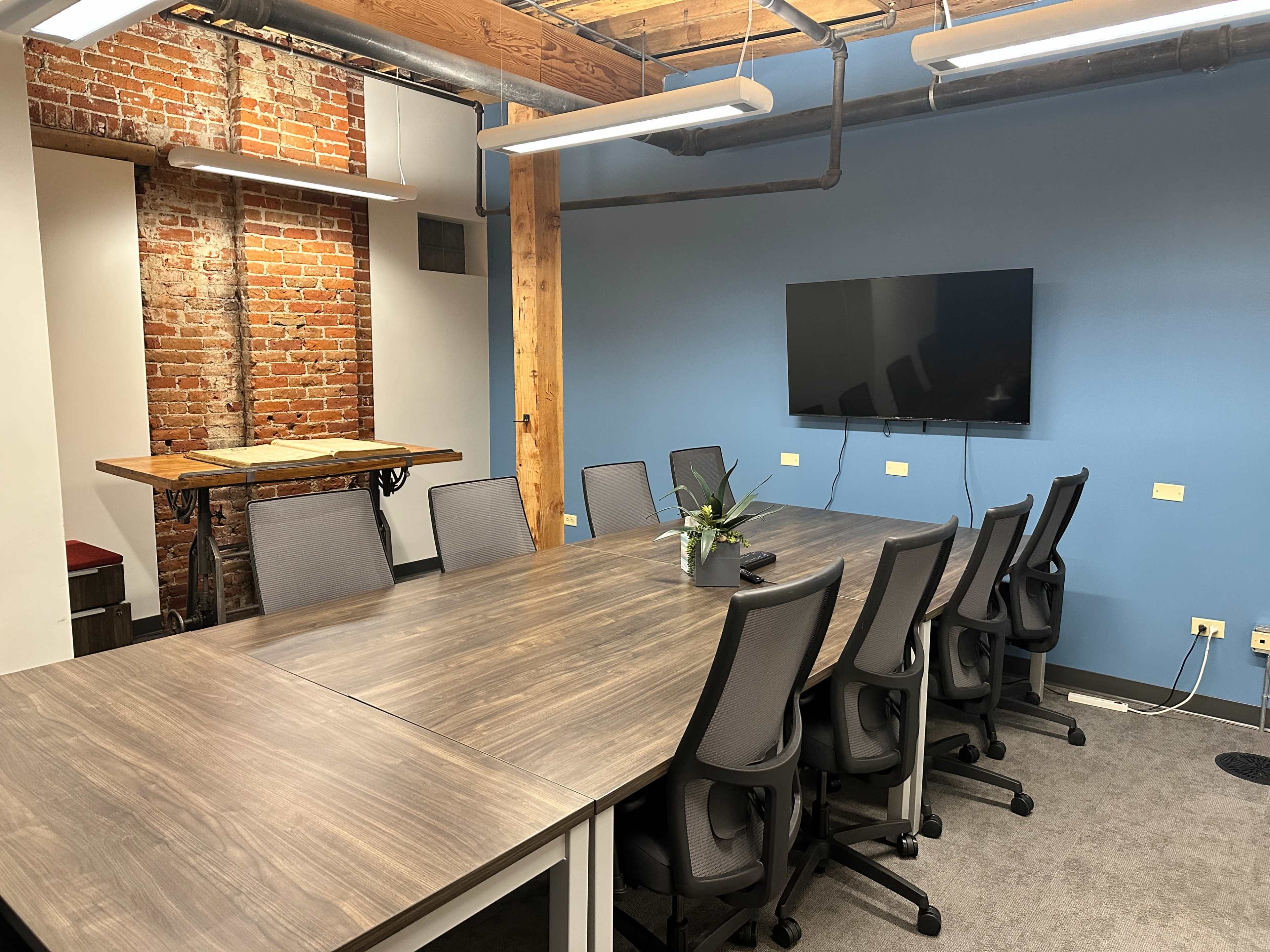 The image shows a modern conference room with a large wooden table, several chairs, a wall-mounted television, and exposed brick walls.
