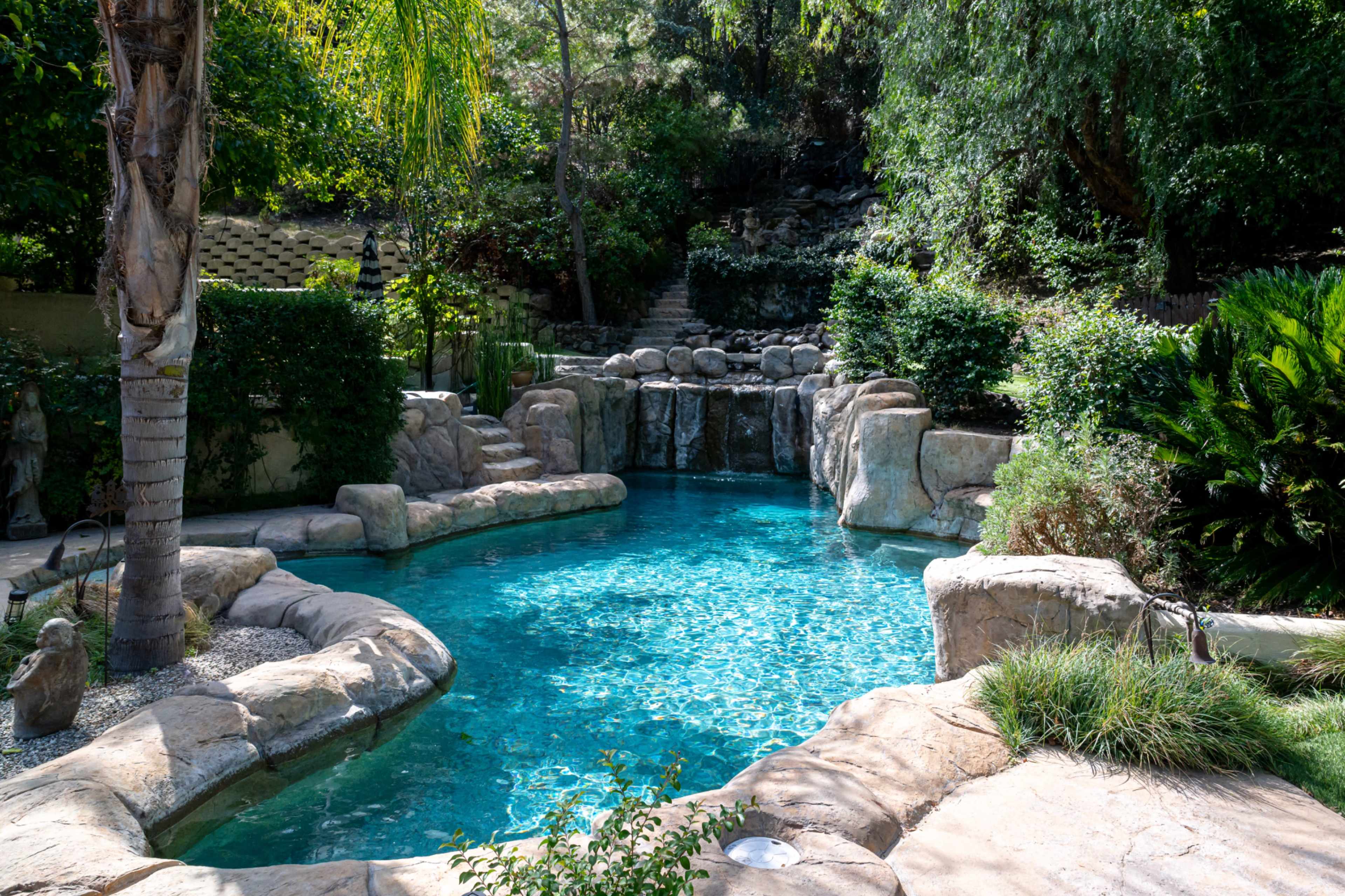 The image shows a landscaped backyard pool featuring natural rock formations and a waterfall, surrounded by lush greenery.