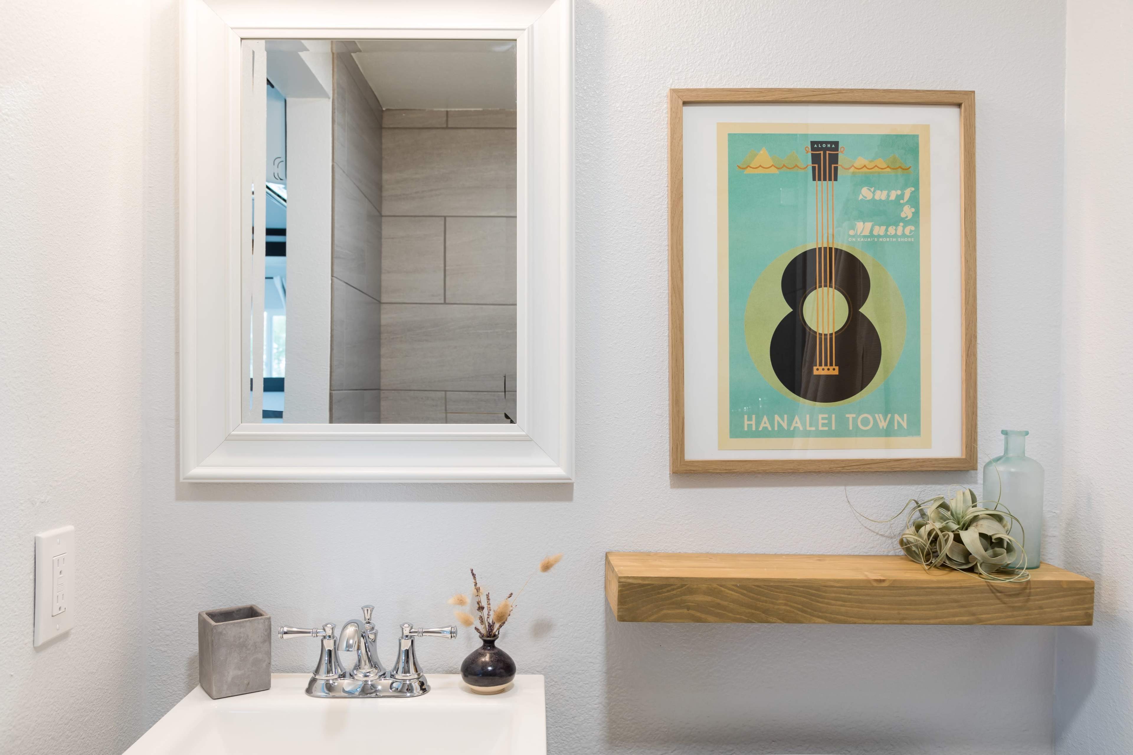 The image shows a clean bathroom with a mirror, a framed poster featuring a guitar, and a wooden shelf displaying decorative items.