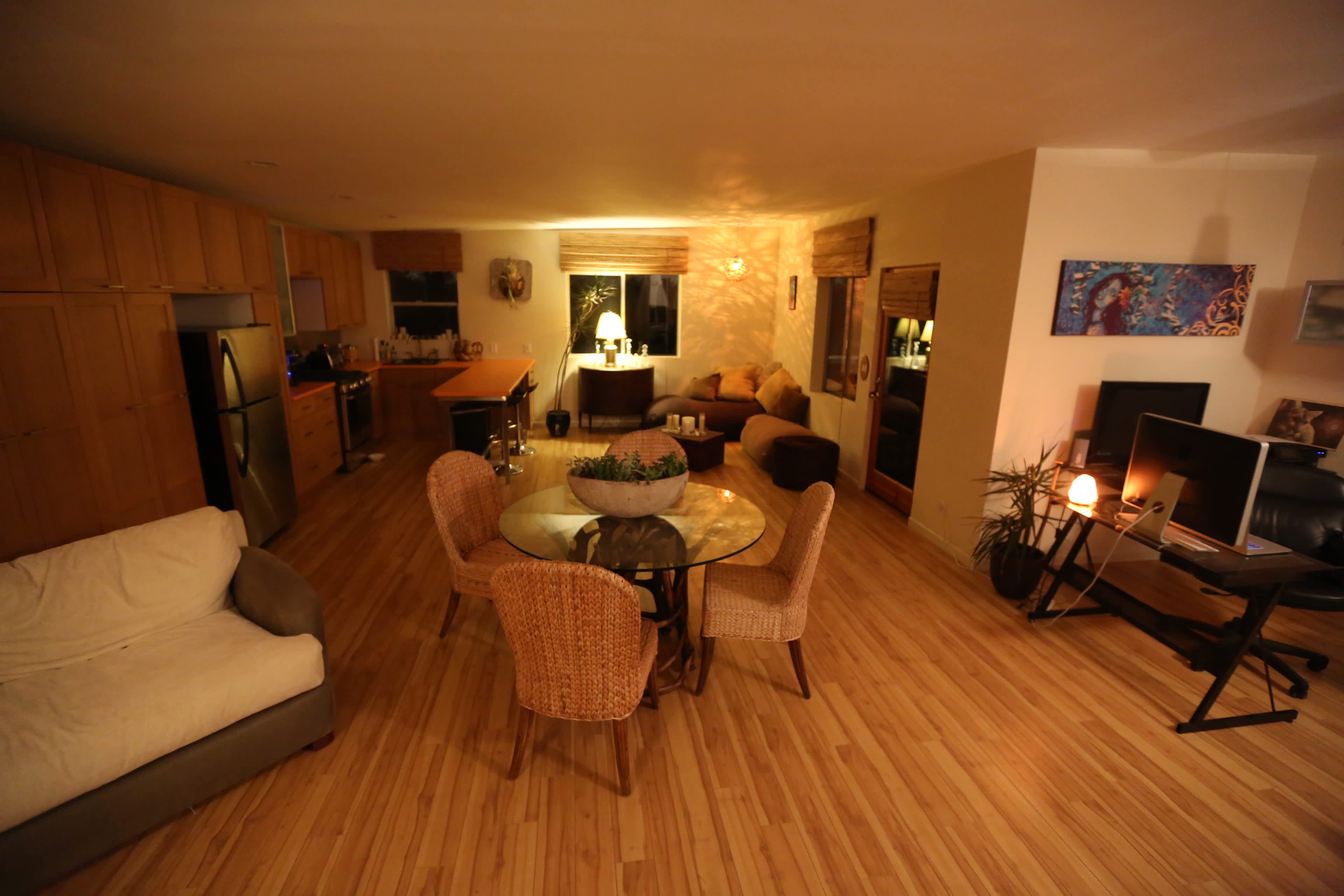 The scene depicts a spacious living area featuring a dining table with chairs, a sofa, and several seating areas, all illuminated by soft lighting.