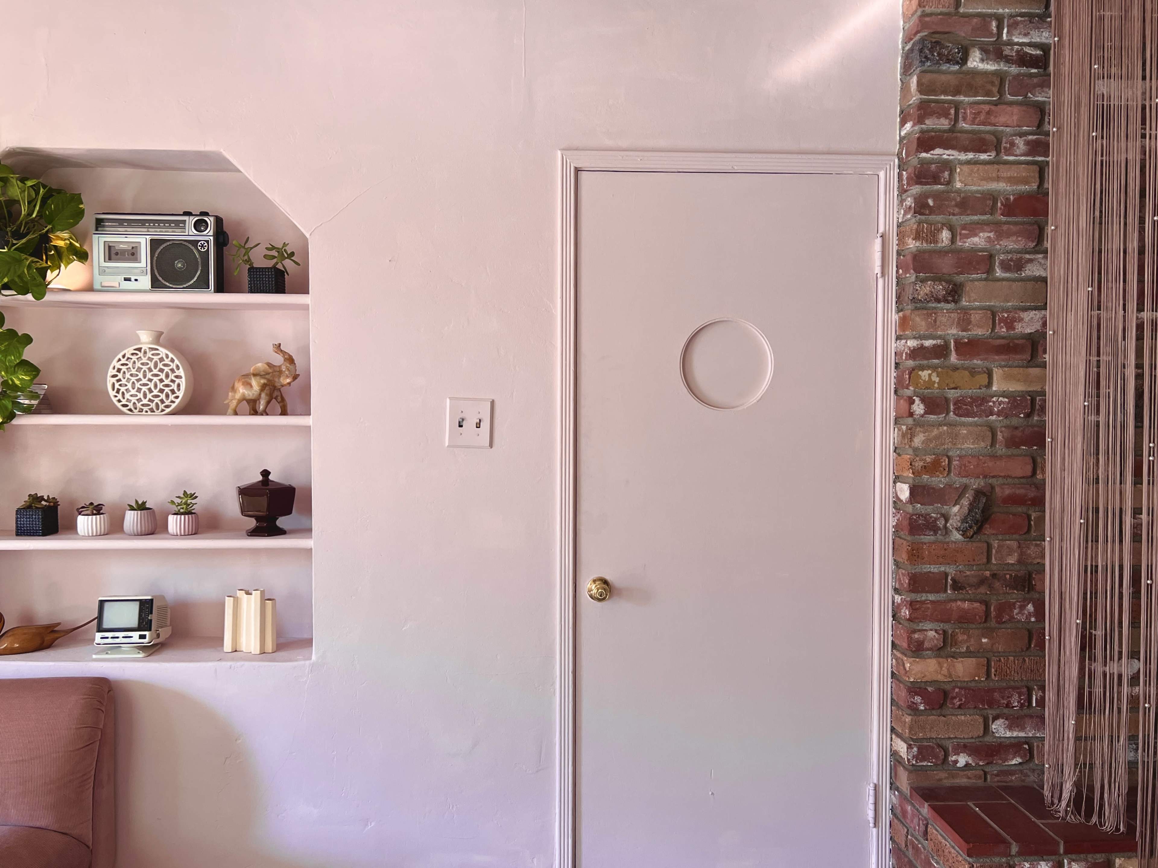 A room with a pink wall, a door with a round window, a brick column, and shelves displaying decorative items and plants.