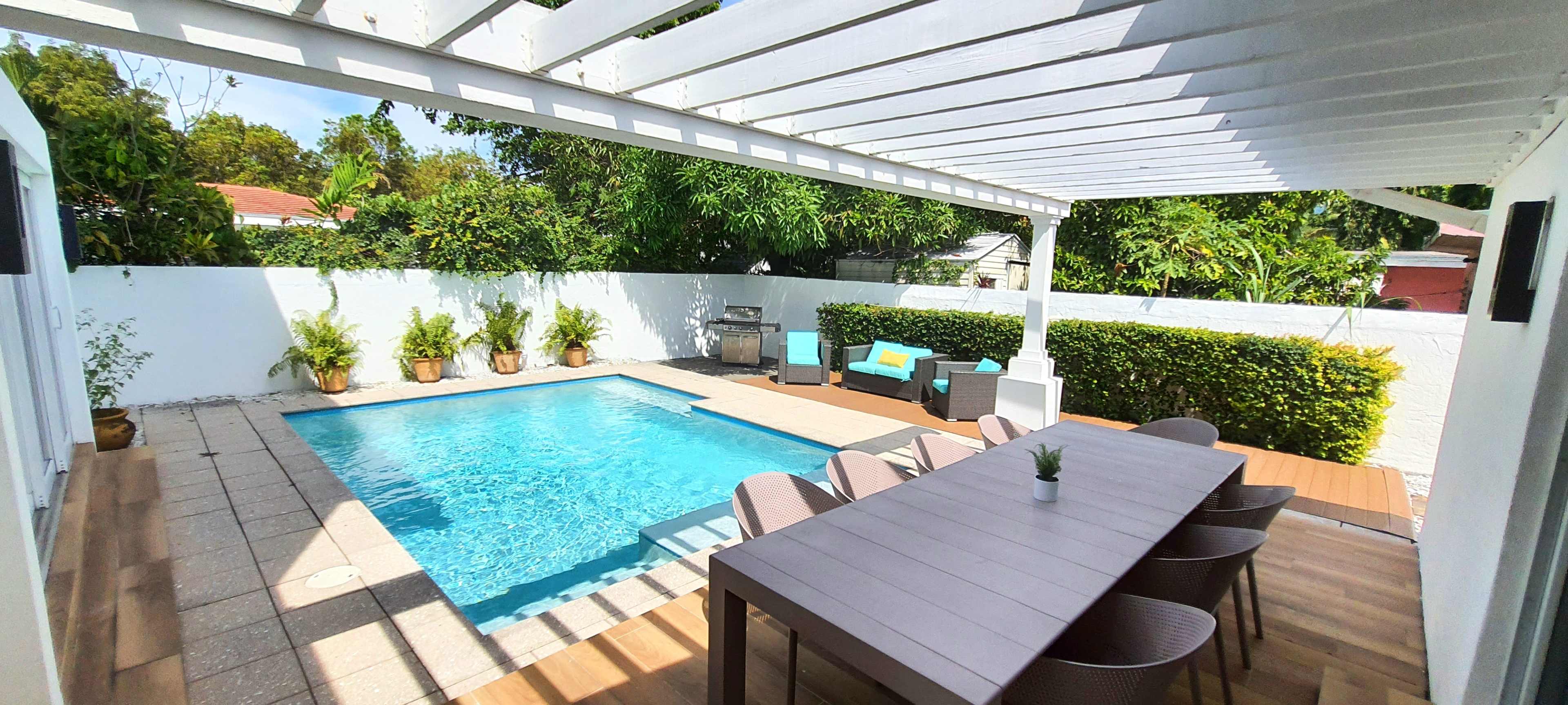 The image shows a backyard patio with a swimming pool, outdoor dining table, and lounge chairs under a covered area.