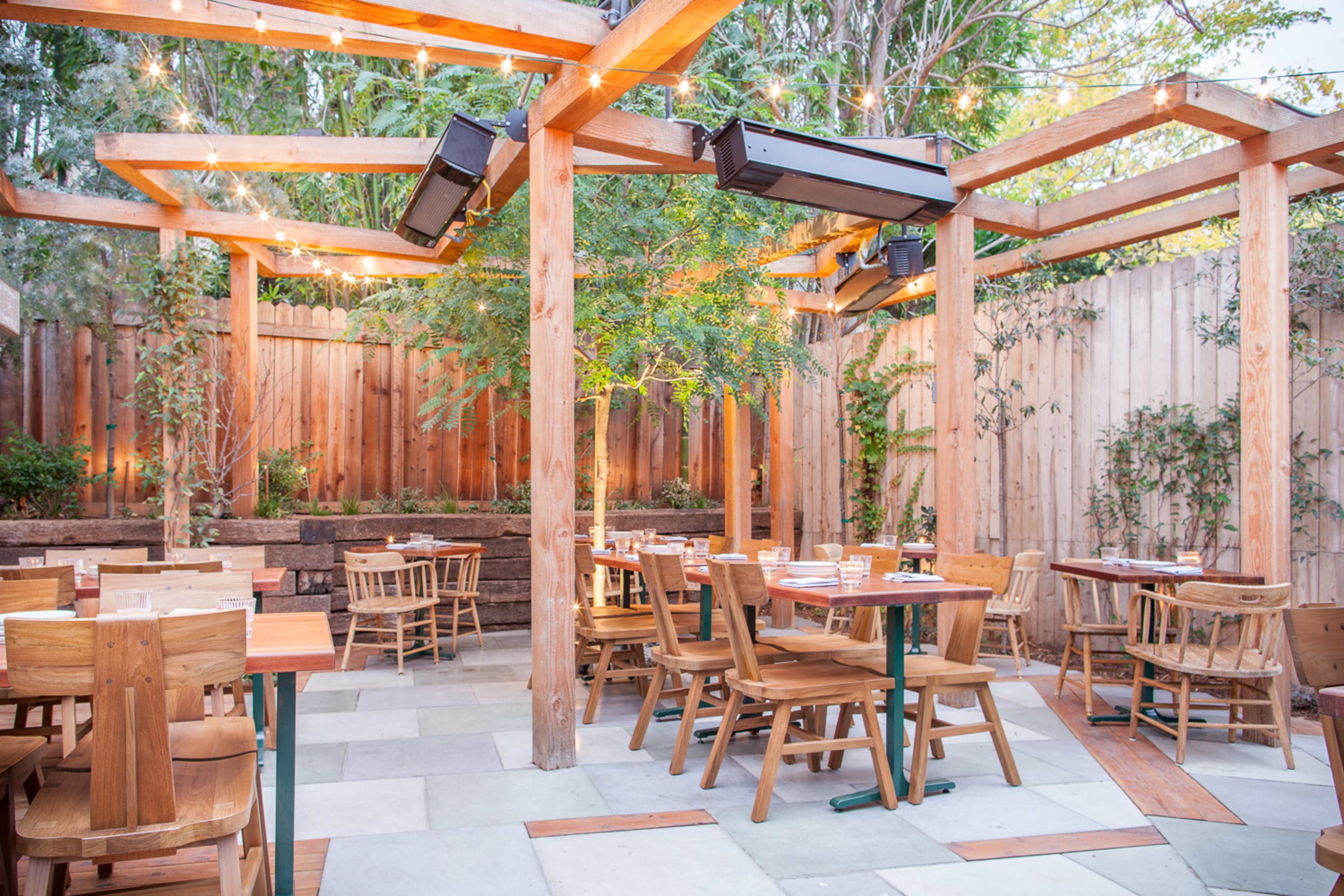 The image shows an outdoor restaurant patio featuring wooden tables and chairs, surrounded by greenery and wooden fencing, with string lights overhead.
