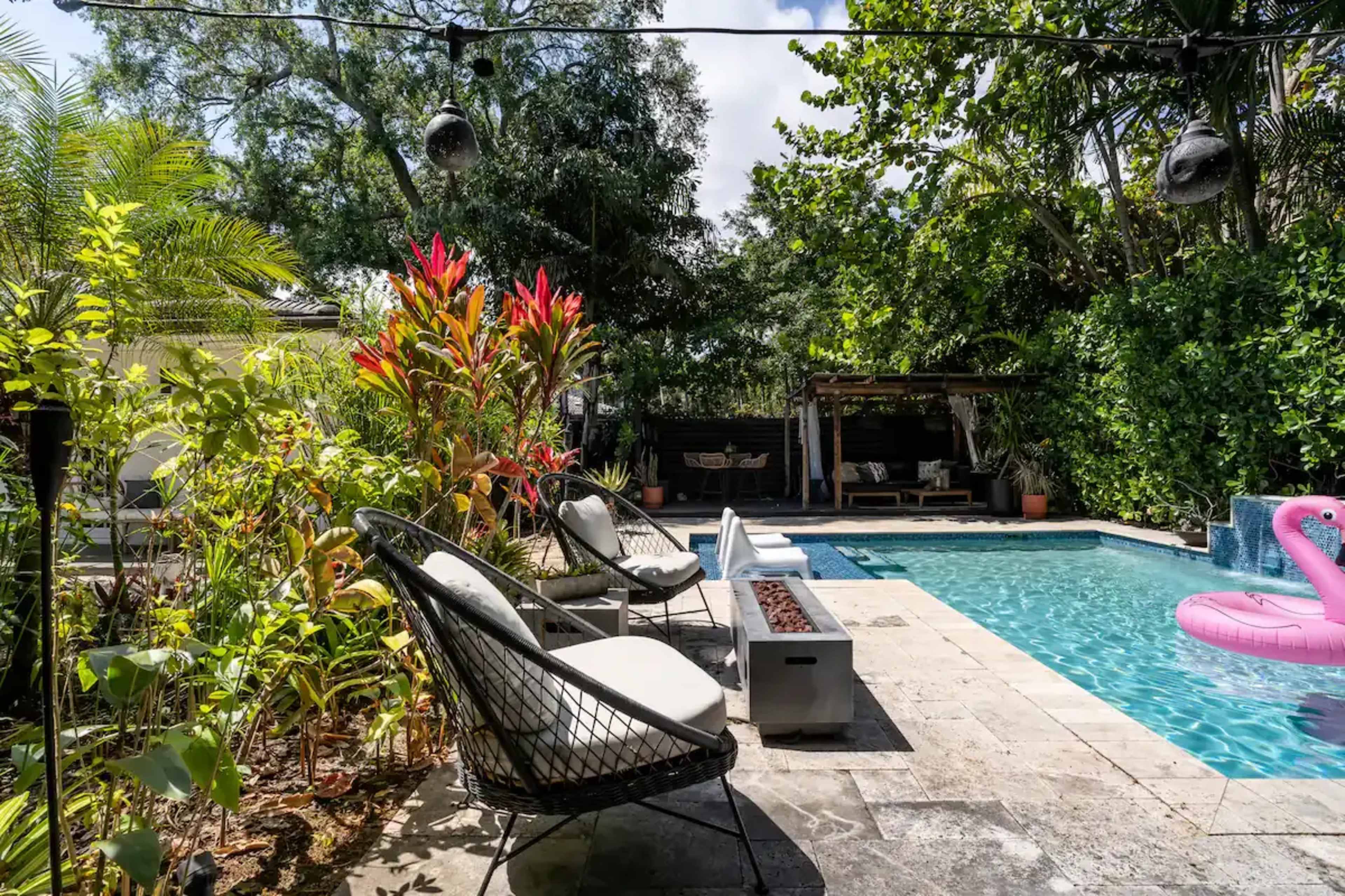 The image shows a backyard with a swimming pool surrounded by lush greenery, lounge chairs, and a tropical landscaping.