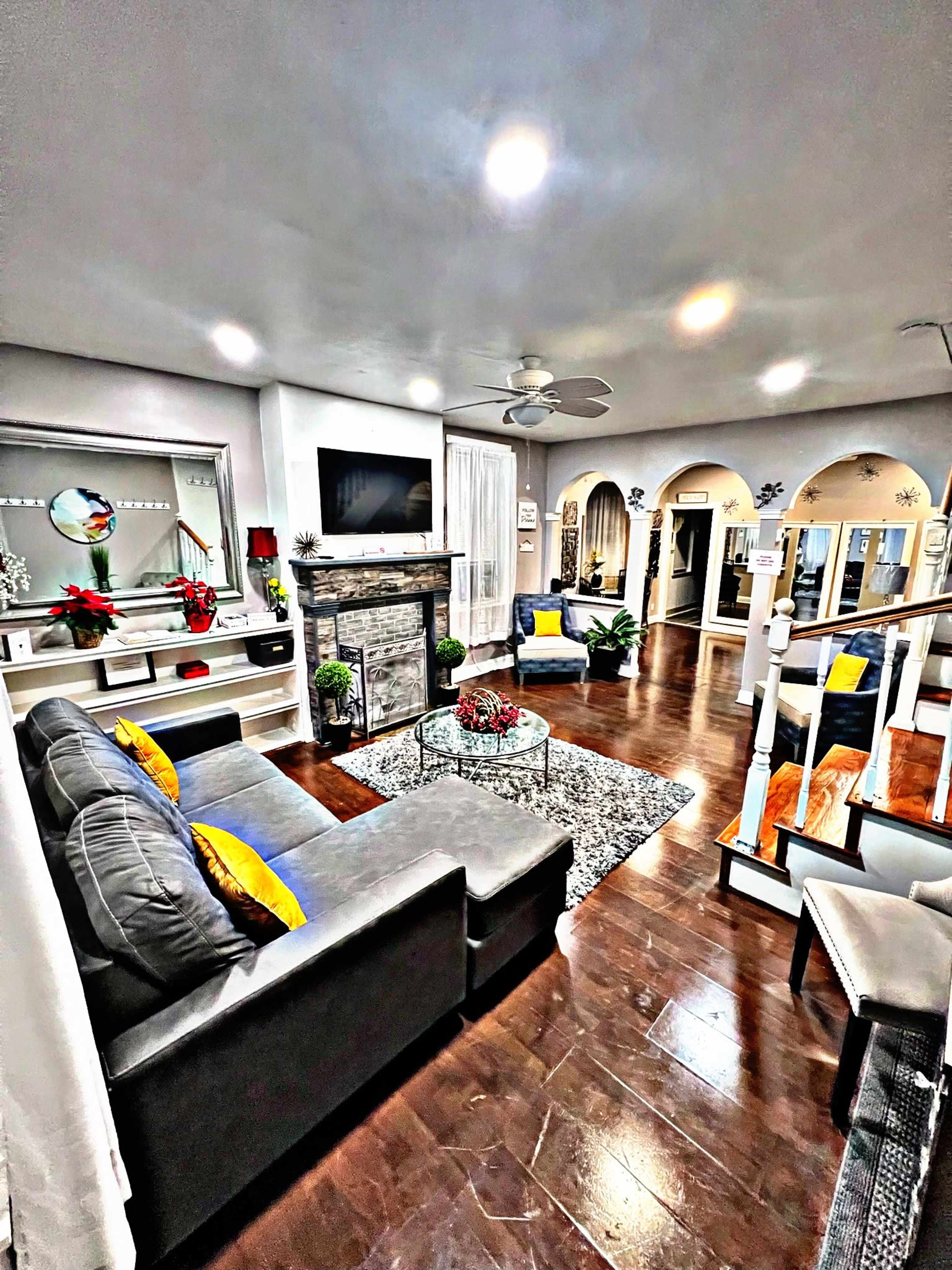 A modern living room features a gray sofa and armchair, a stone fireplace, and a glass coffee table, all arranged under bright ceiling lights.