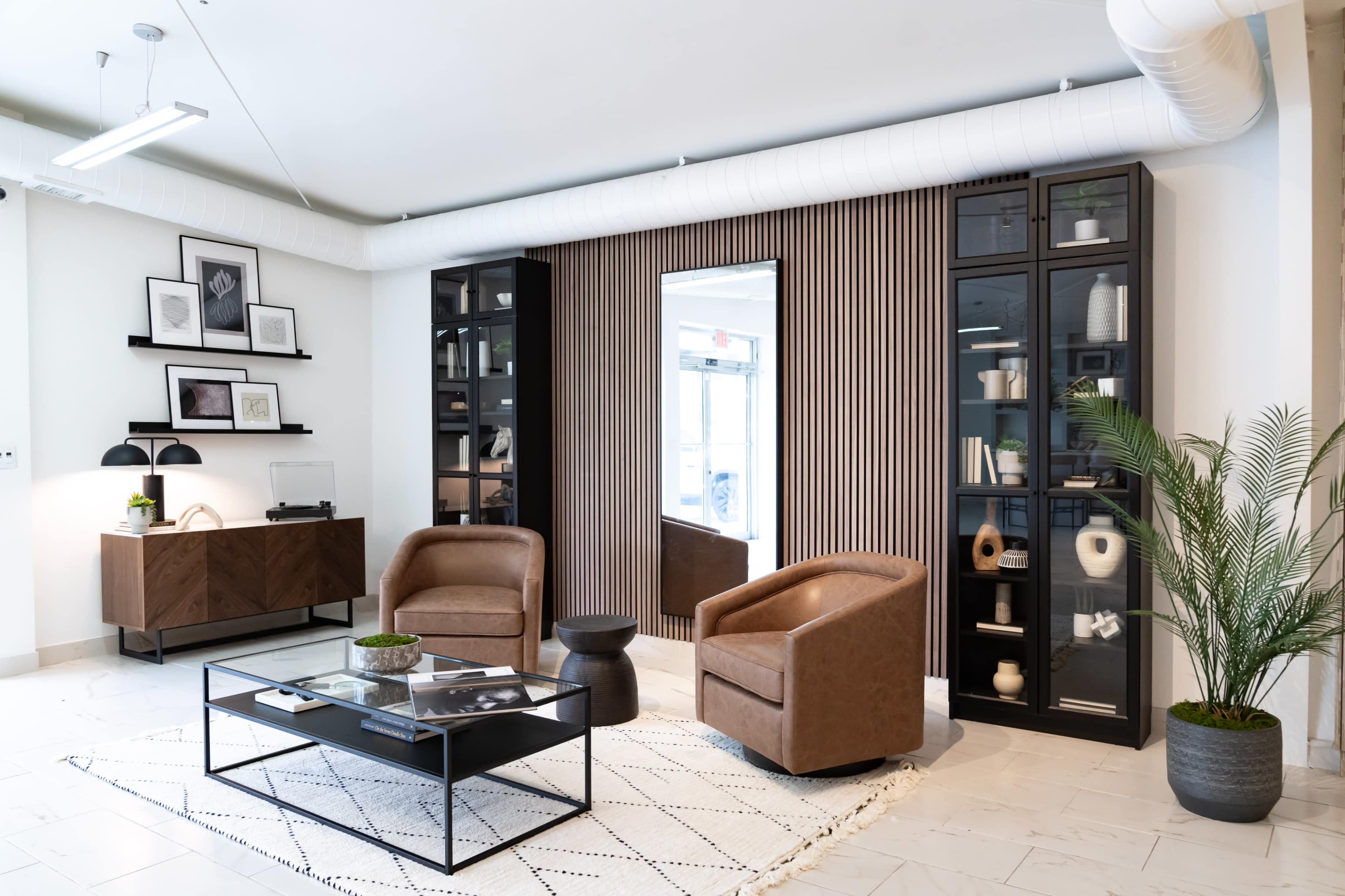 The image shows a modern living room set with two brown leather chairs, a glass coffee table, a patterned rug, and shelves displaying decorative items against a wall with vertical wood paneling.