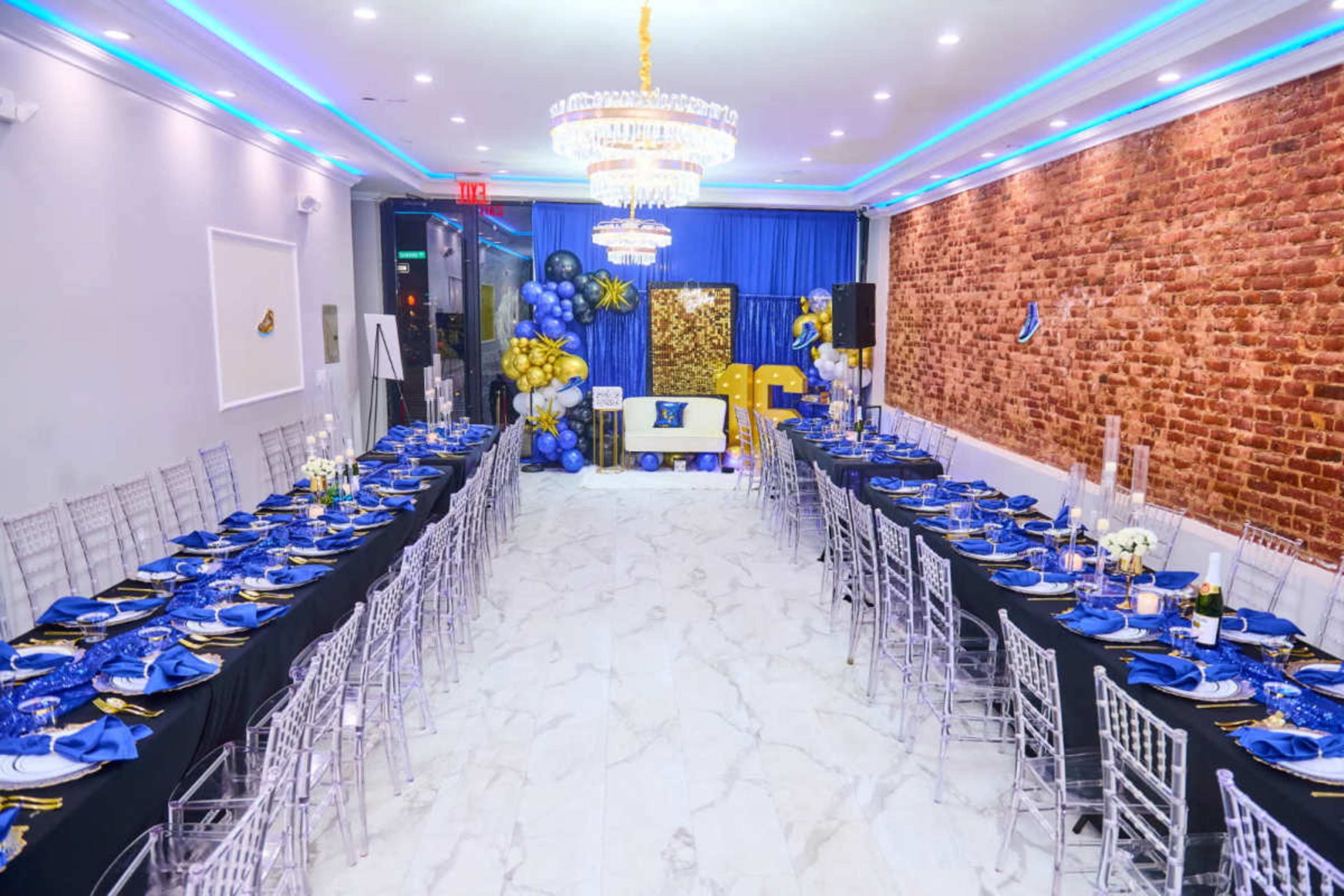 The image shows a decorated indoor event space with two long tables set with blue and gold tableware, flanked by balloons and a brick wall backdrop.