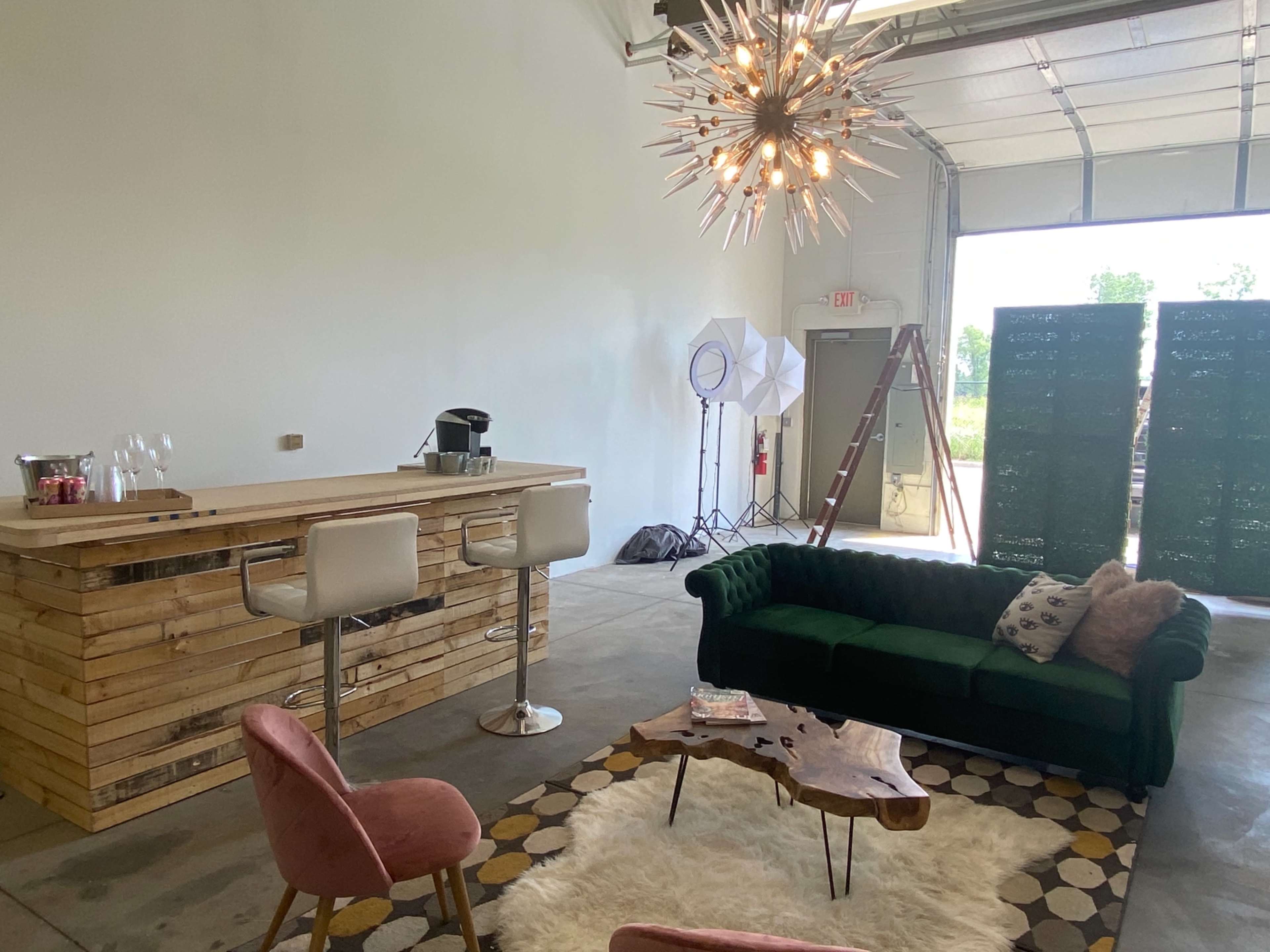 A modern interior space features a wooden bar with stools, a green sofa, pink chairs, a coffee table, and a large light fixture, with an open garage door in the background.