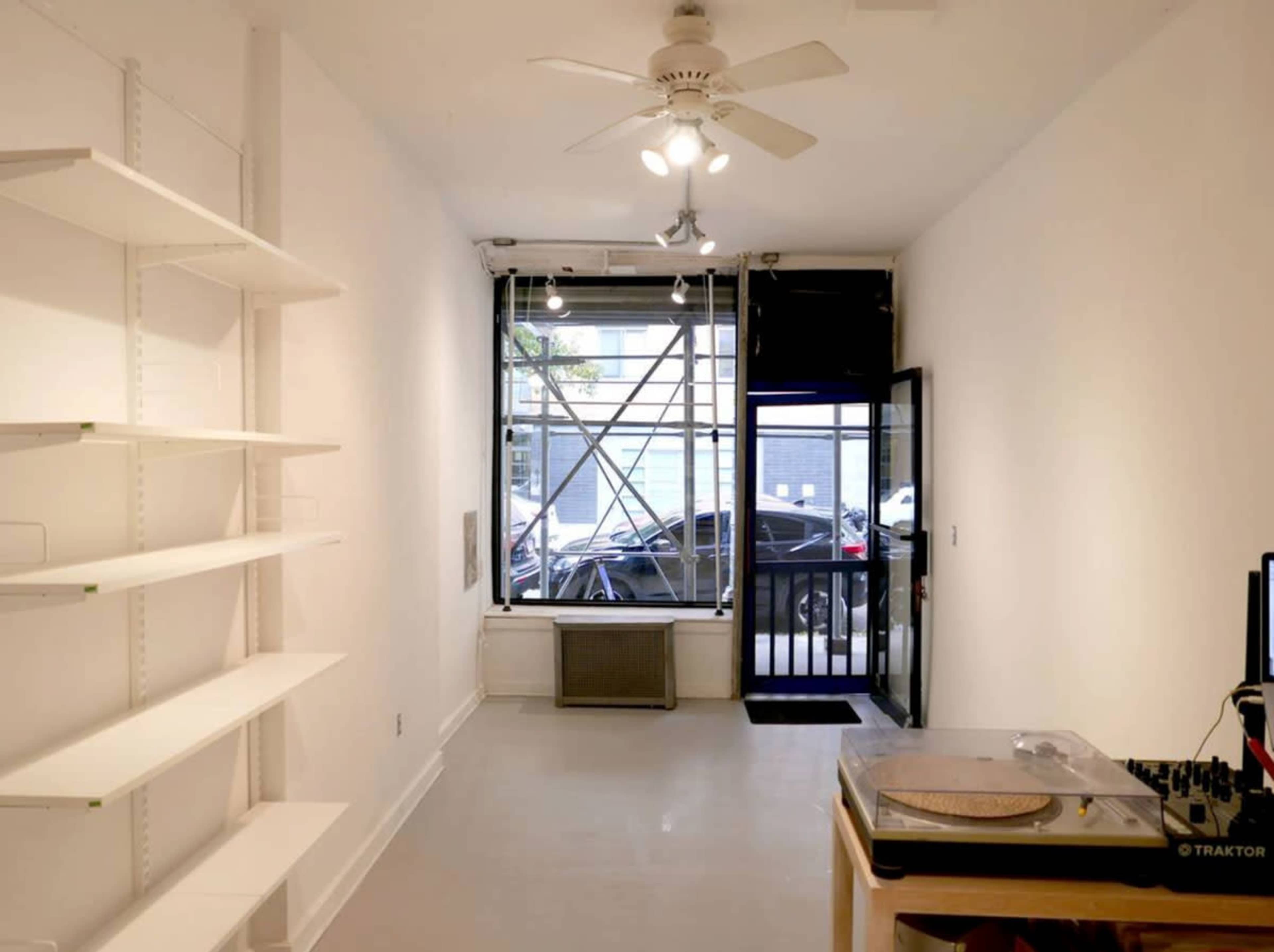 A minimally furnished room with a ceiling fan, open entrance, and shelves along one wall.