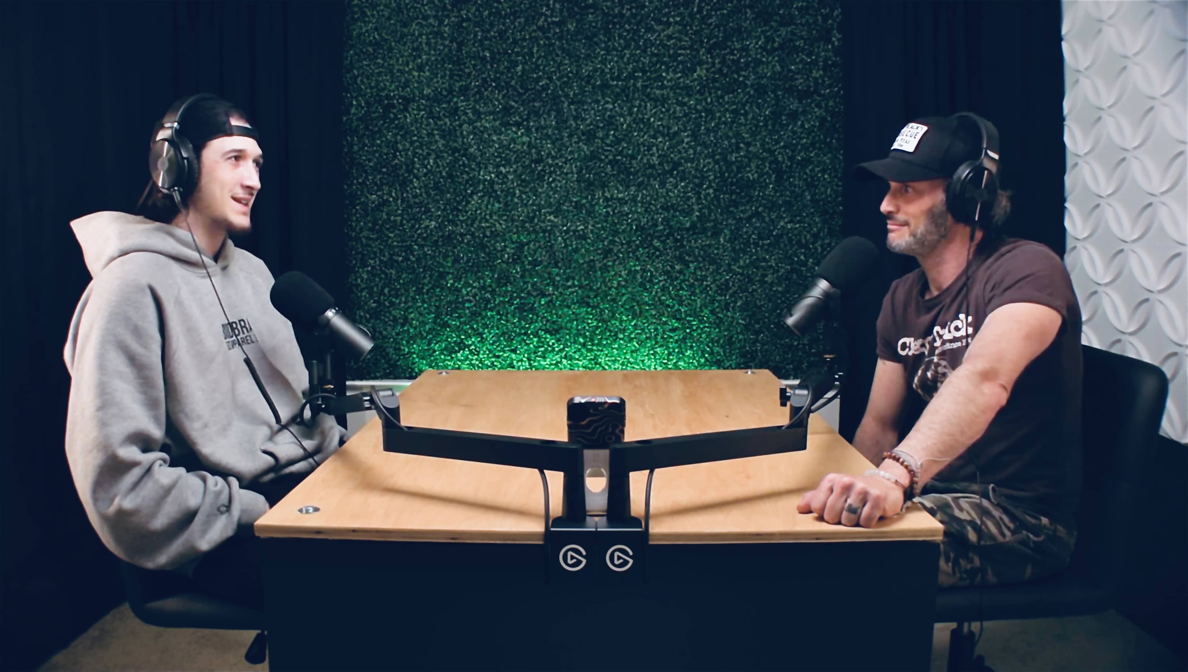 Two individuals sit at a table with microphones, engaged in conversation against a wall featuring greenery.