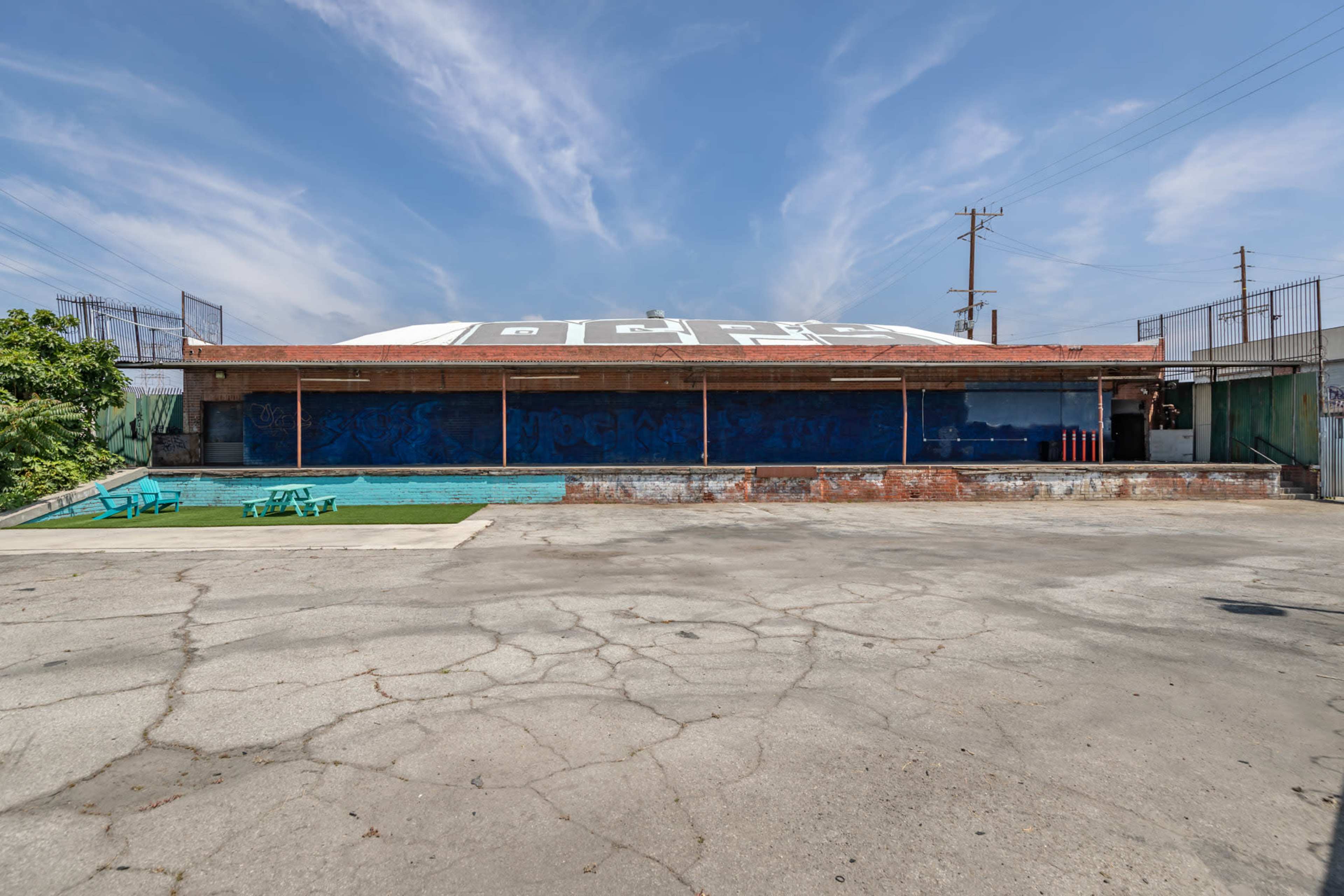 The image shows an empty outdoor area with a faded, mural-covered wall, a pool, and blue seating against a clear sky.
