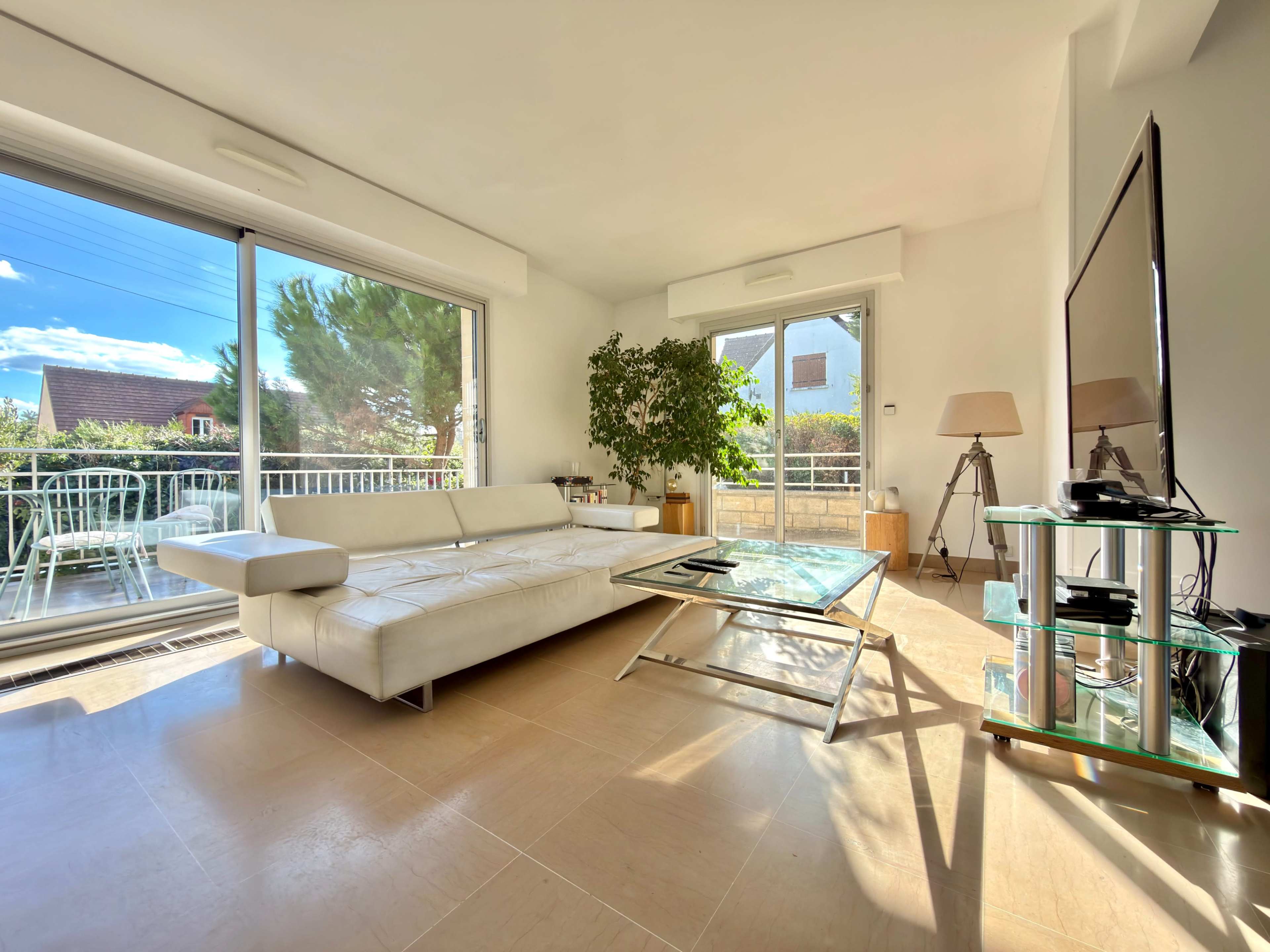 The image shows a bright living room with a white sectional sofa, a glass coffee table, and a television on a glass stand, alongside large windows that open to a balcony.