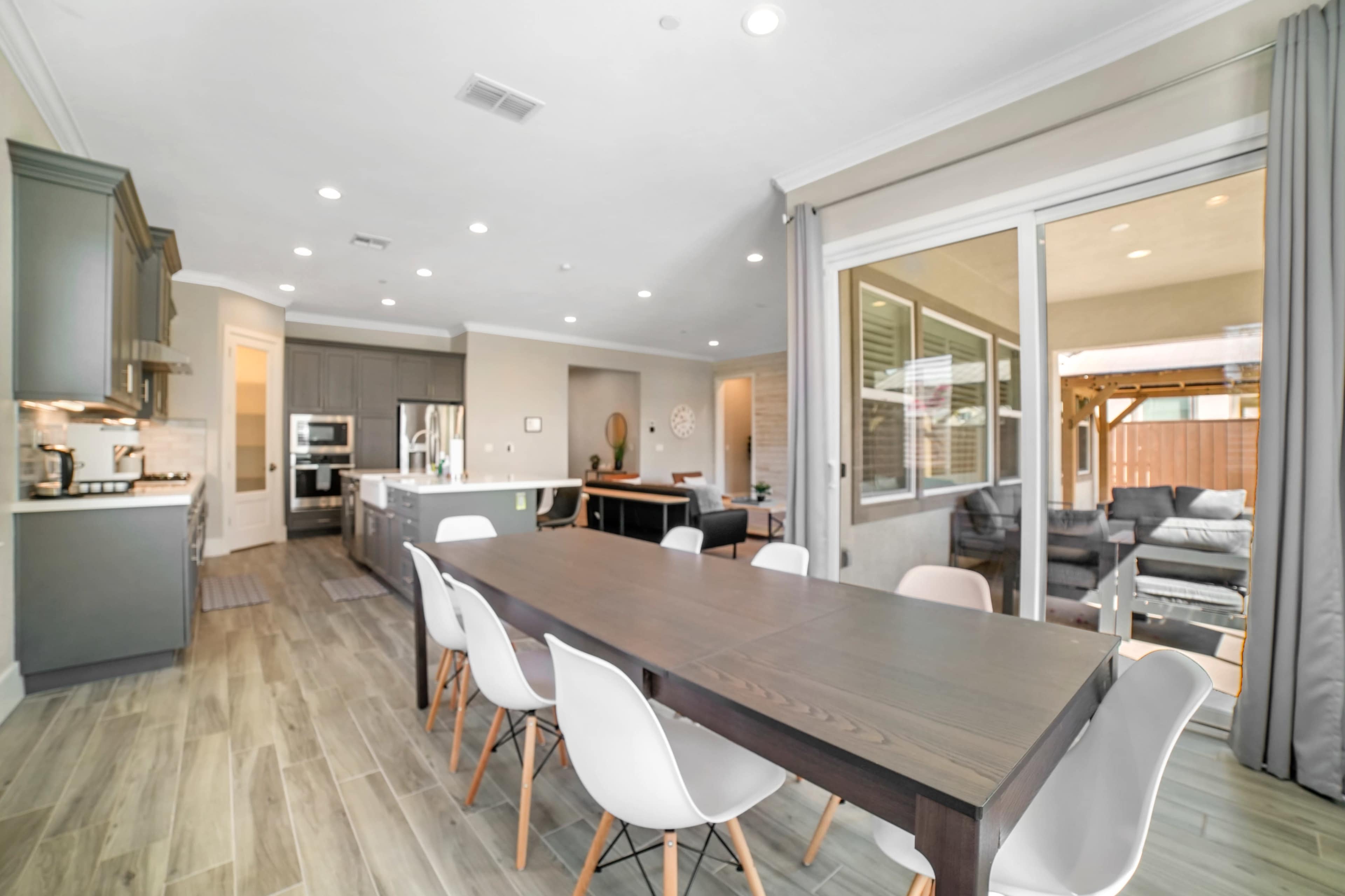 A modern kitchen and dining area feature a long wooden table surrounded by white chairs, with large windows providing natural light and views of an outdoor seating space.