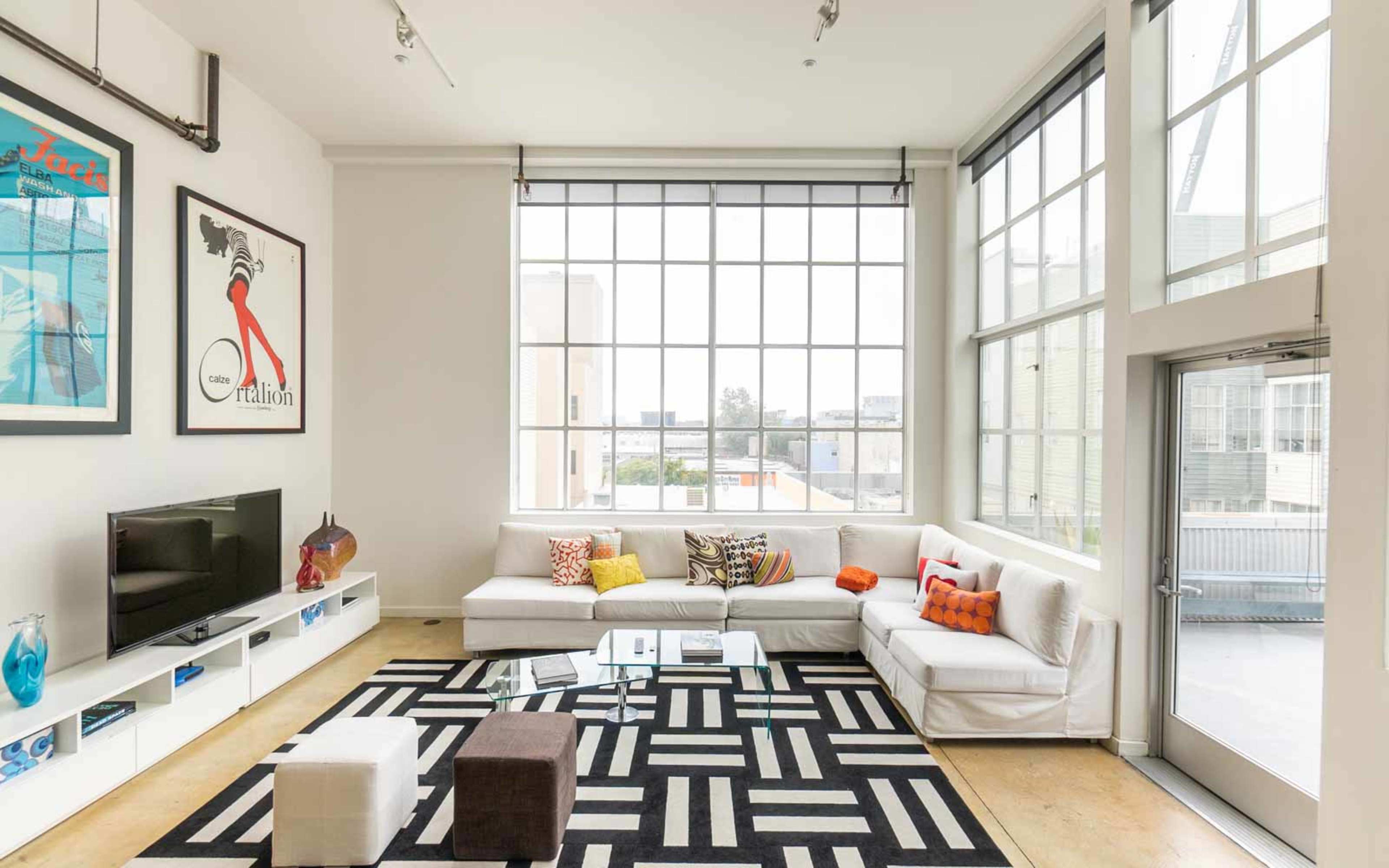 A modern living room with large windows, a white sectional sofa, black and white patterned rug, and a television on a minimalist shelf.