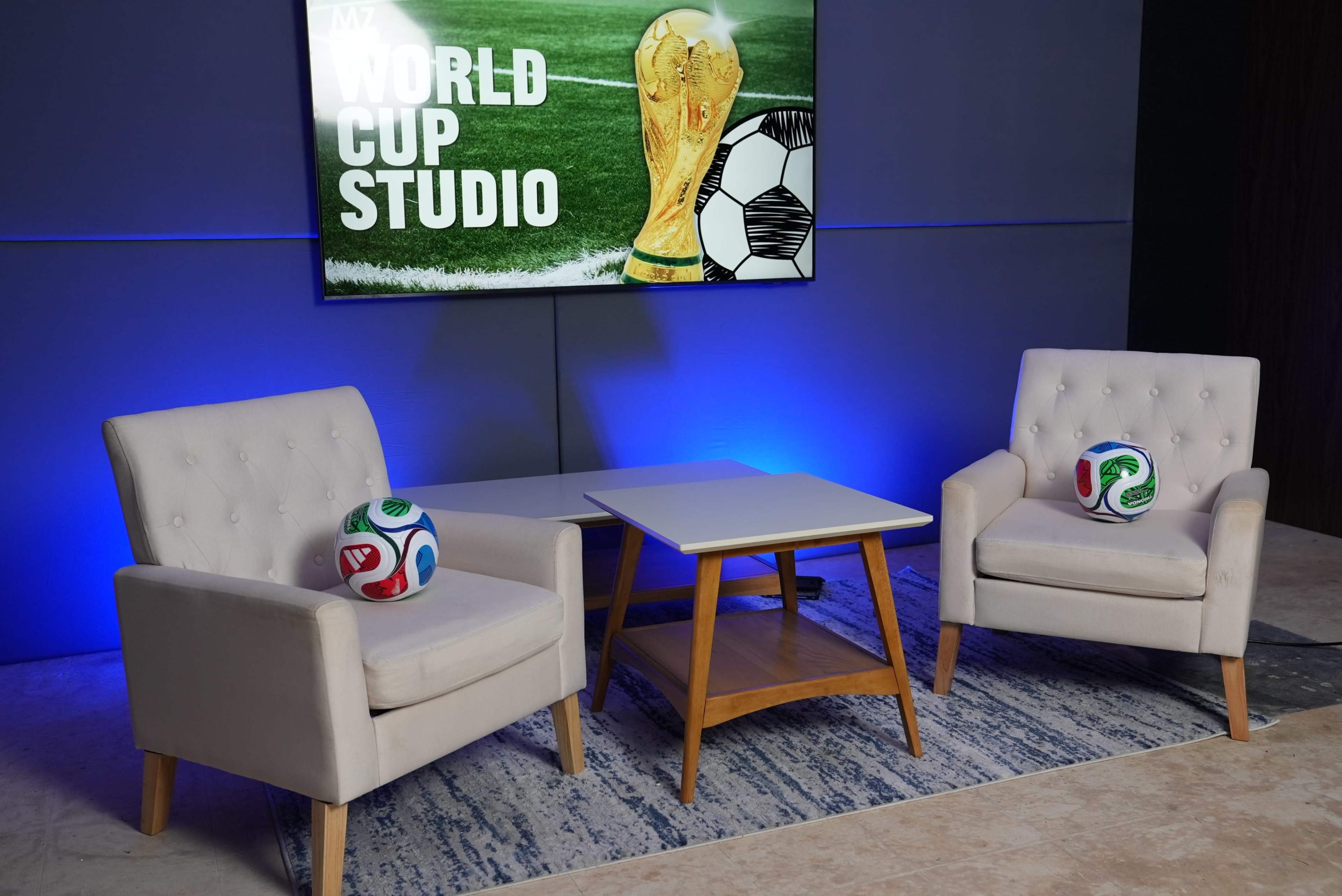 The image shows a studio setup for a World Cup segment, featuring two white armchairs, a small wooden table, and decorative soccer balls on the floor in front of a screen displaying the World Cup trophy and branding.