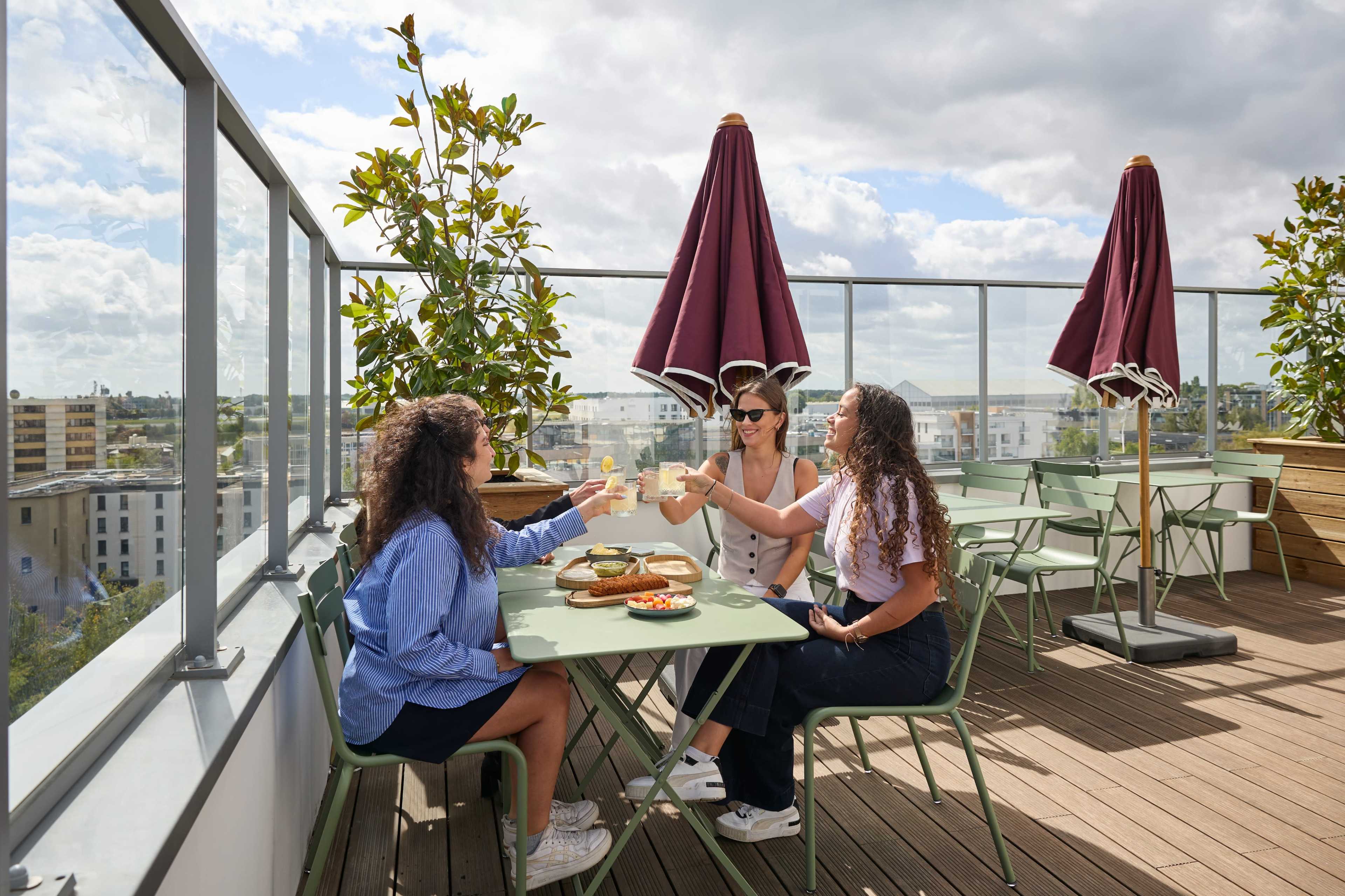 Three women are seated at a rooftop table, toasting with drinks while enjoying a meal under shaded umbrellas.