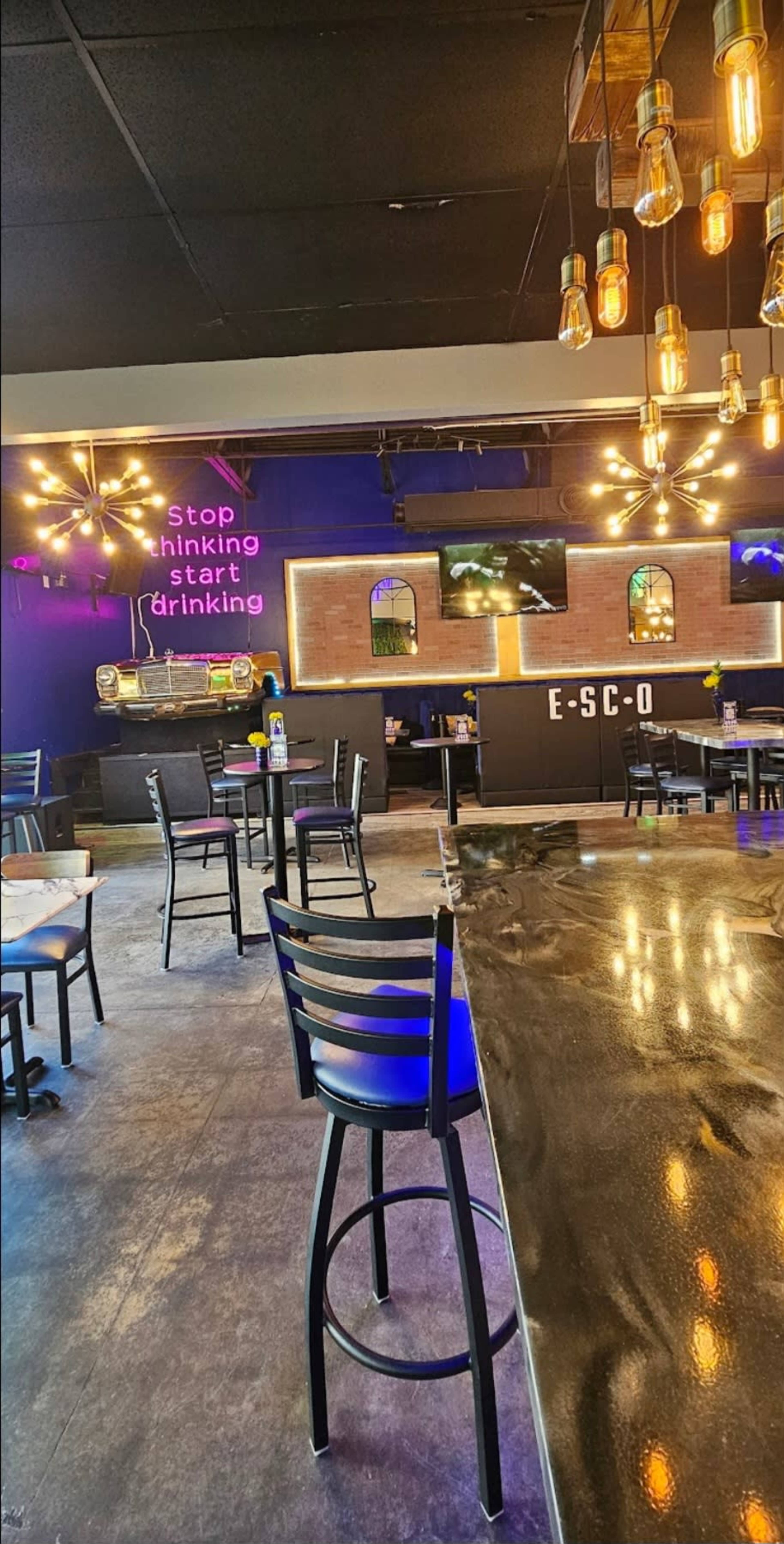 The image shows a modern bar interior with black tables and chairs, illuminated by hanging lights, featuring a neon sign that reads "stop thinking start drinking."