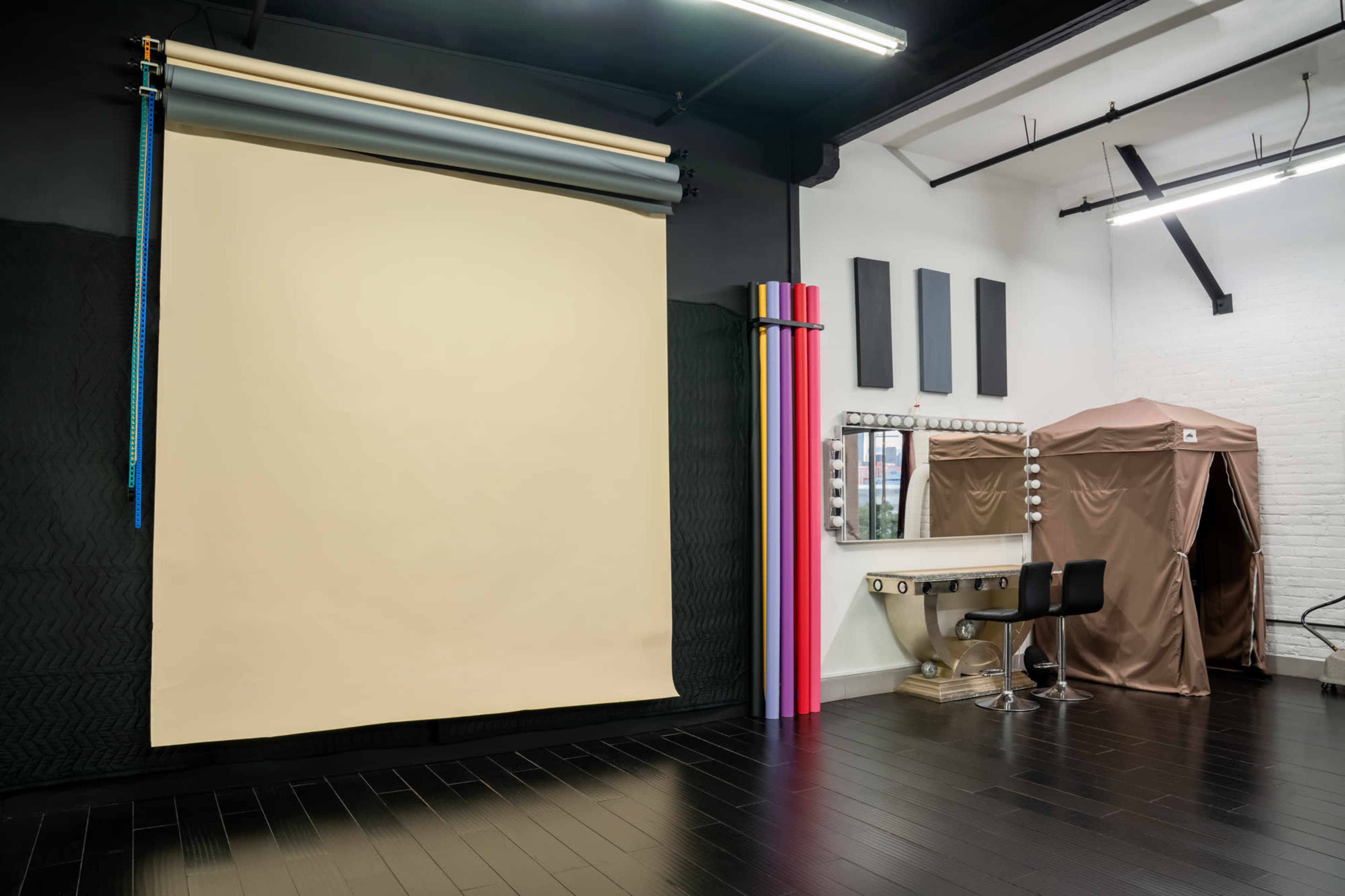 The image shows a photography studio with a beige backdrop, makeup station with a mirror and lights, and a cozy changing area.