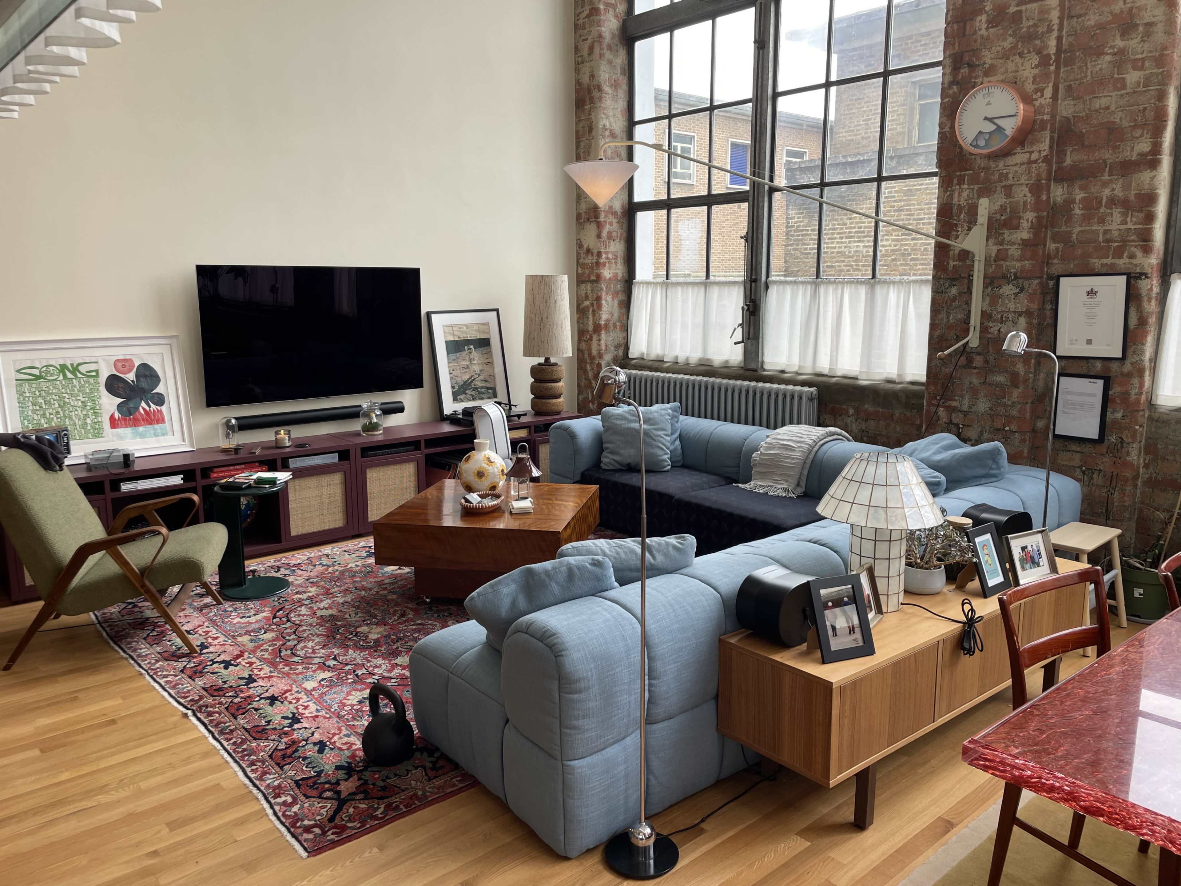 The image shows a stylish living room featuring blue sofas, a large television, a wooden coffee table, and a mix of decorative items along with large windows letting in natural light.