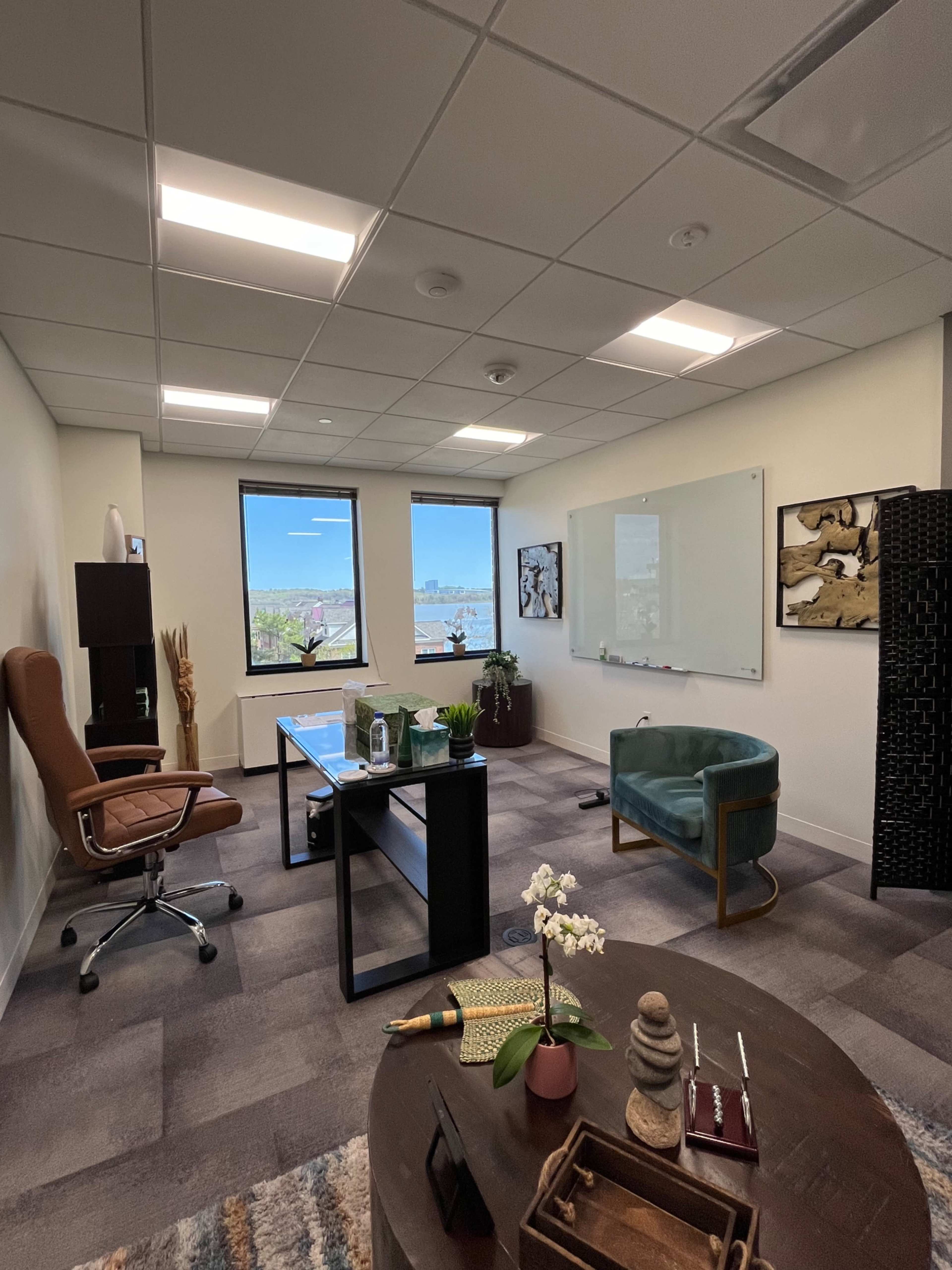 A modern office space with a wooden desk, ergonomic chair, potted plants, and a whiteboard on one wall, overlooking a scenic view through large windows.
