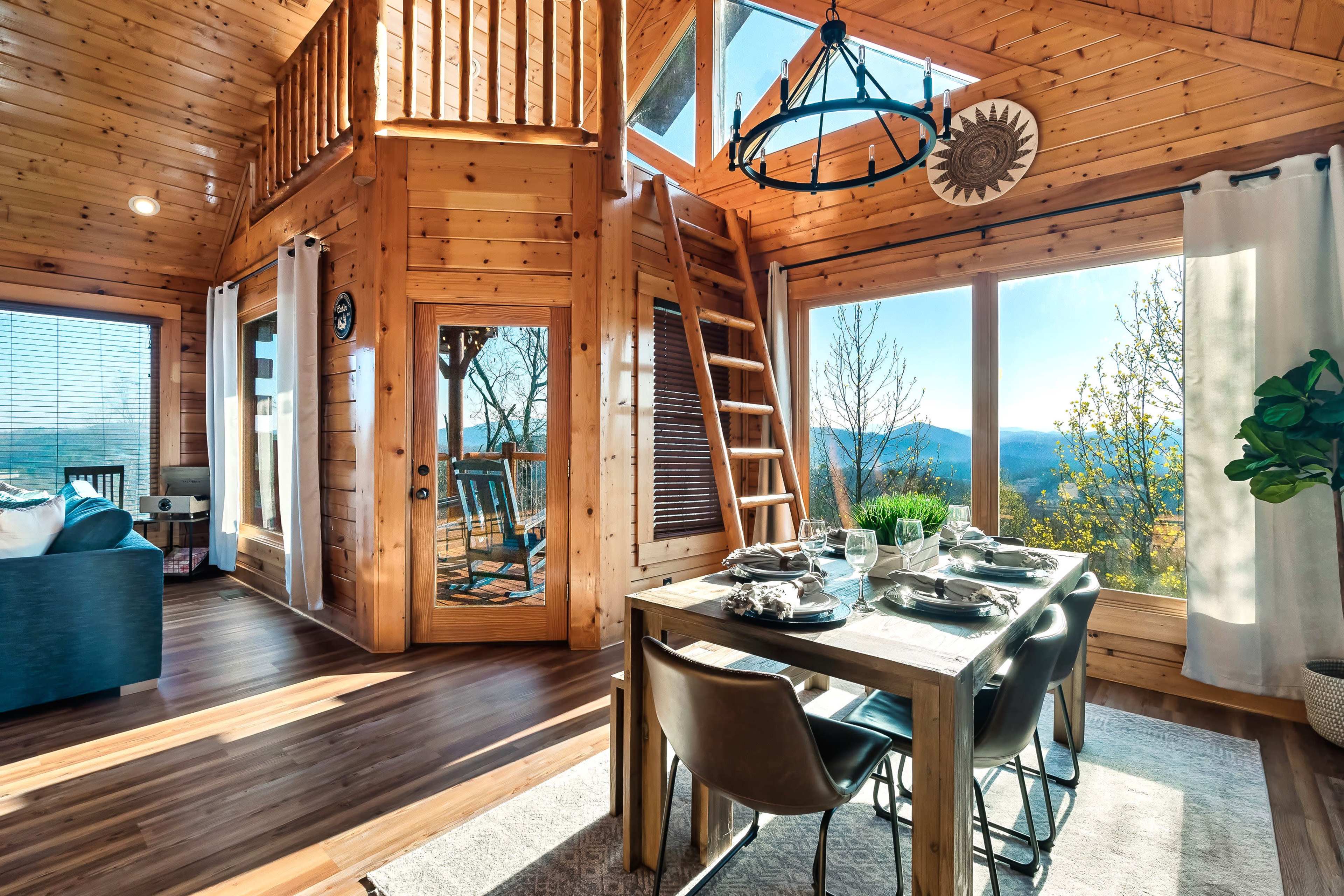The image shows a spacious wooden interior of a cabin with a dining area, a staircase leading to a loft, large windows offering mountain views, and a cozy seating arrangement.