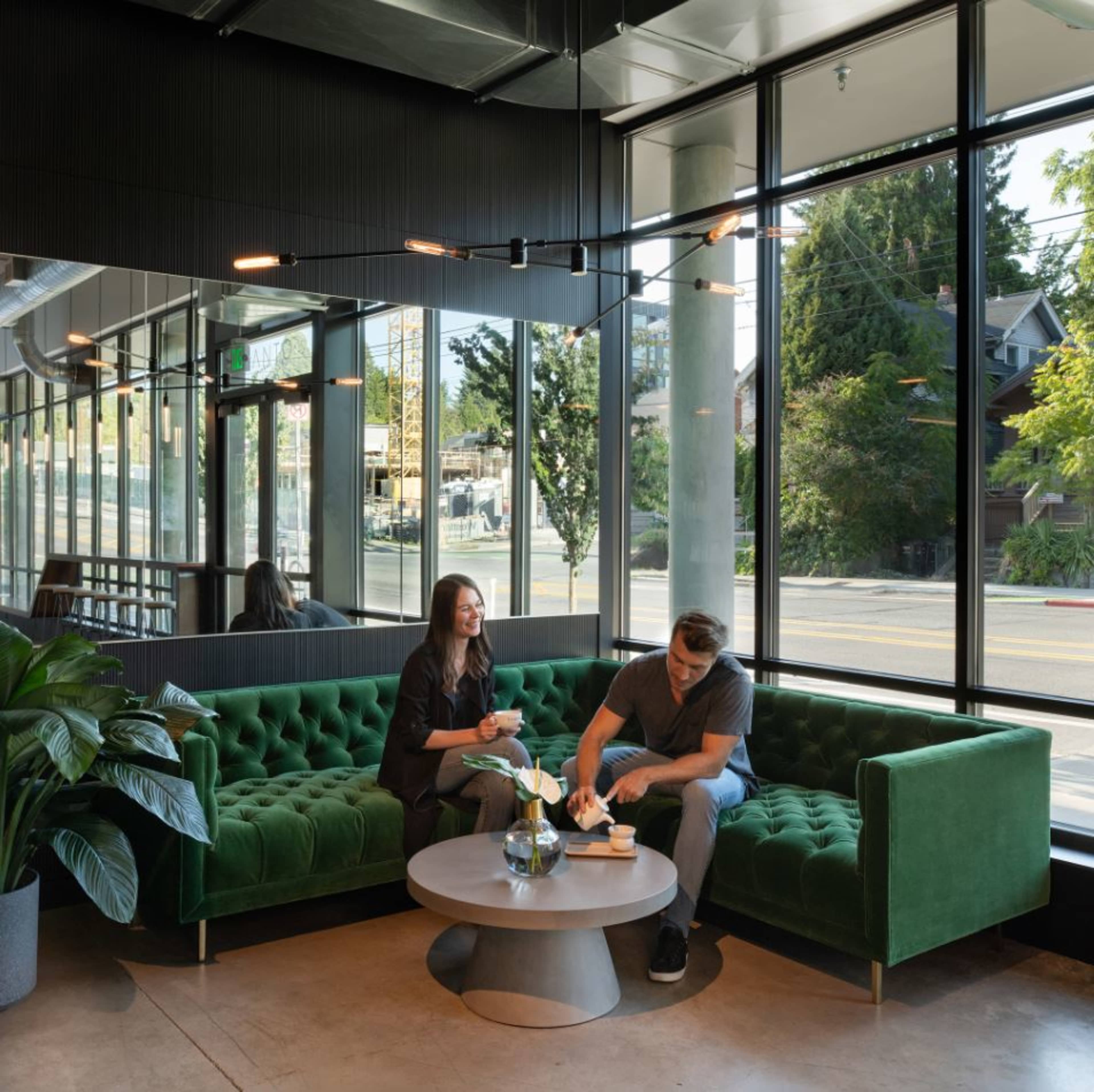 A man and woman sit on a green tufted sofa in a modern cafe with large windows, surrounded by plants and a round table.