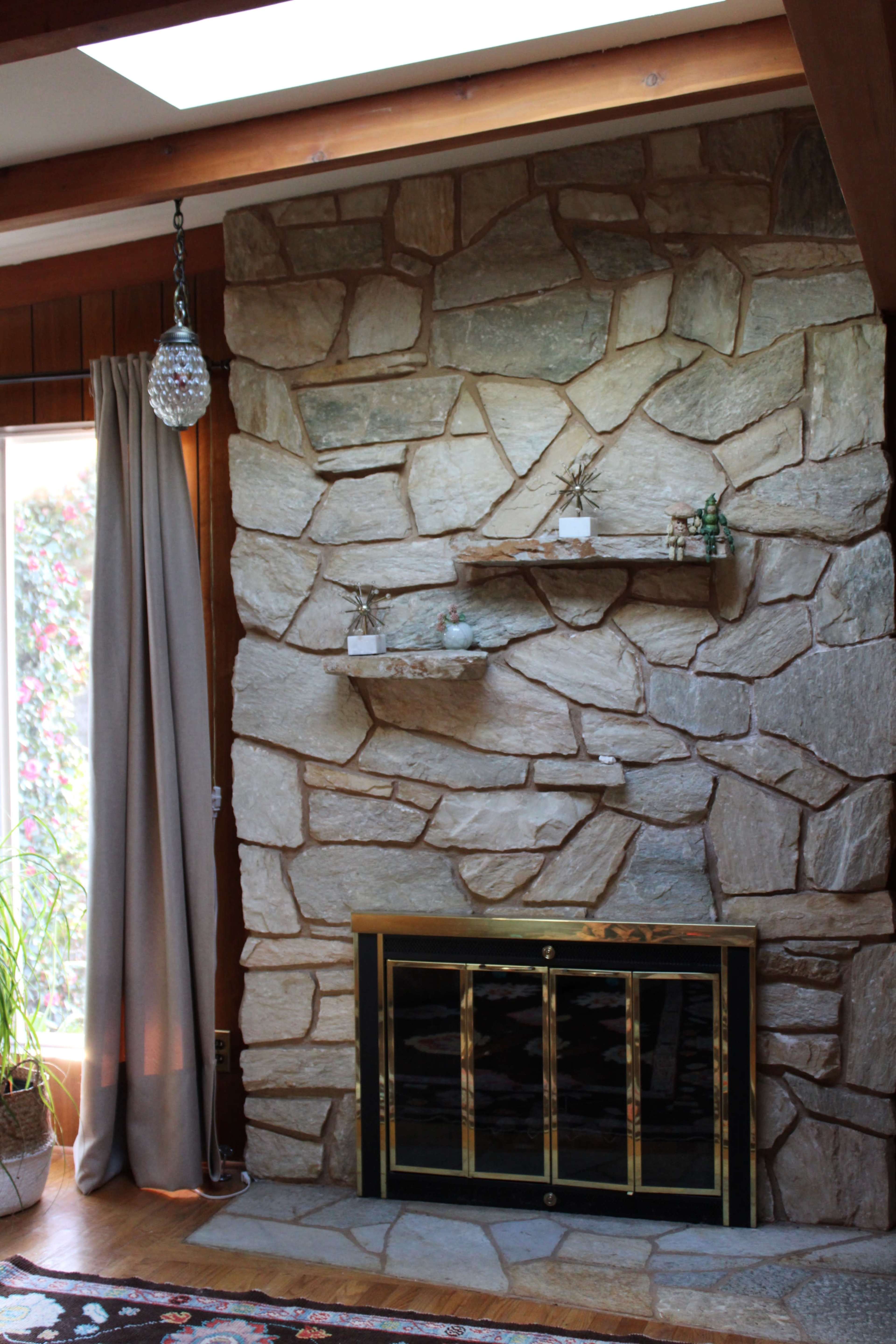 The image shows a stone fireplace with a gold-framed glass door, accompanied by a couple of decorative shelves and a nearby window draped with a curtain.
