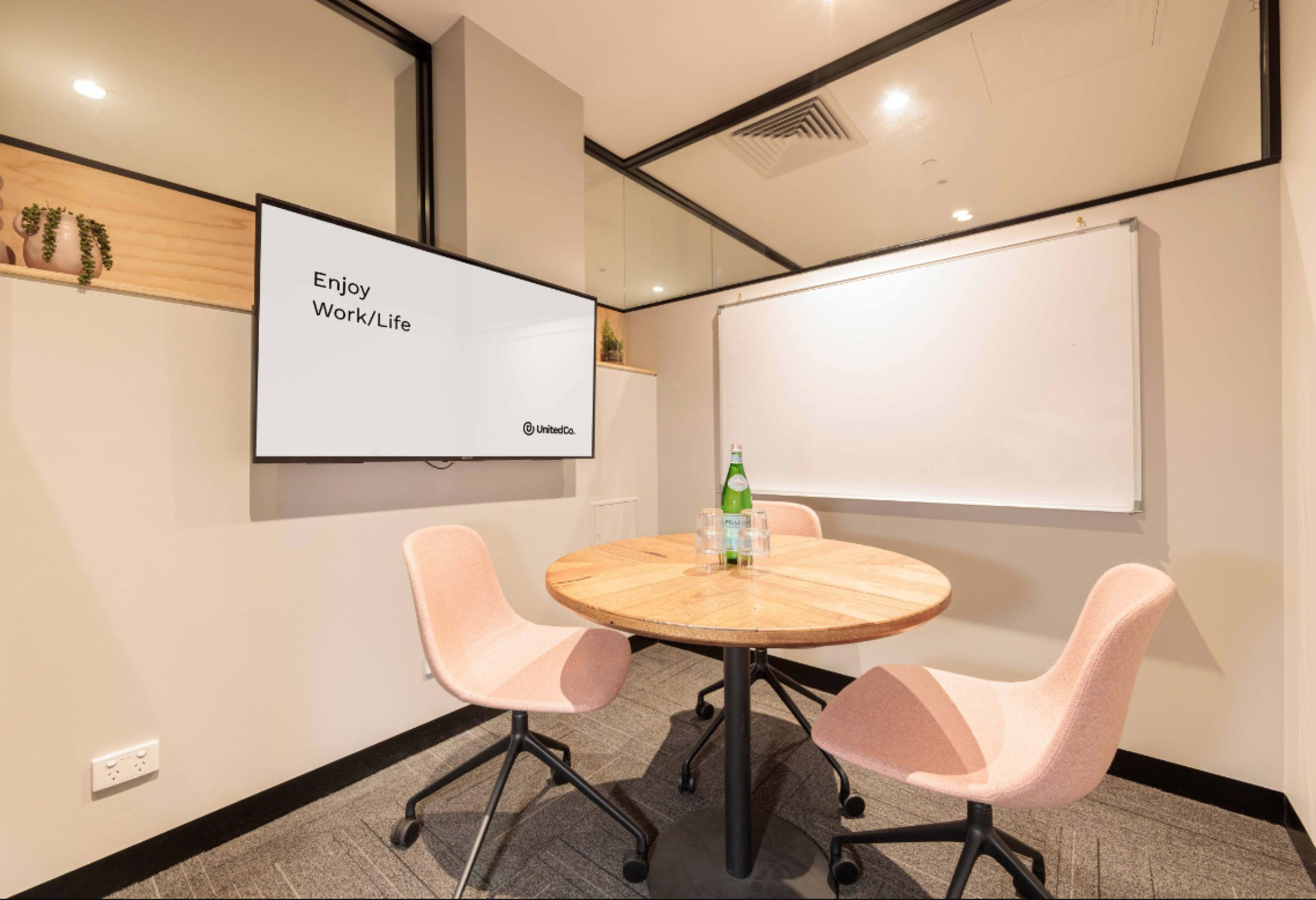 A small meeting room features a round wooden table with two pink chairs, a screen displaying "Enjoy Work/Life," and a whiteboard on the wall.