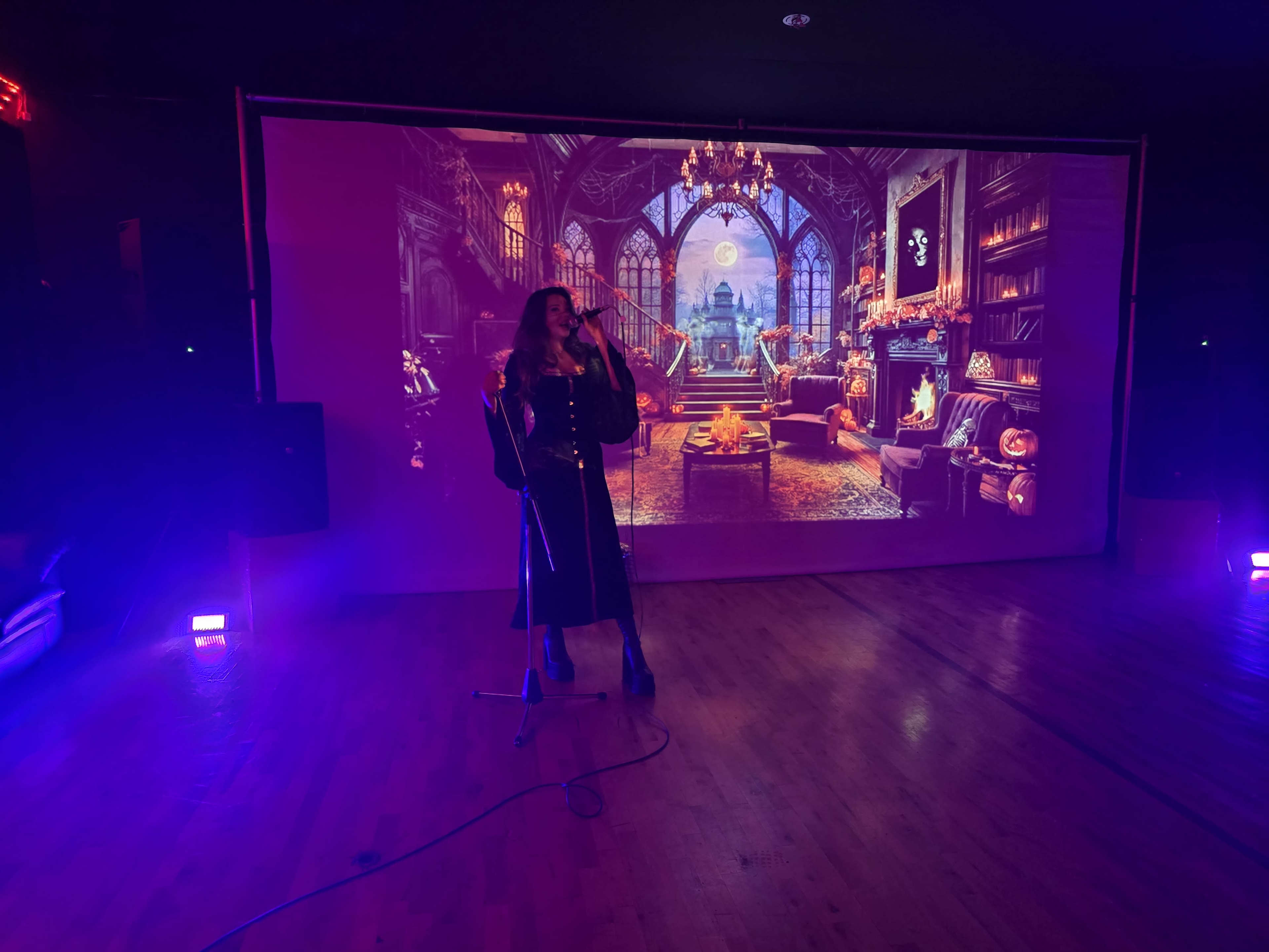 A woman stands on a stage with a microphone in front of a large projection depicting a cozy, ornate room with a fireplace and large windows.