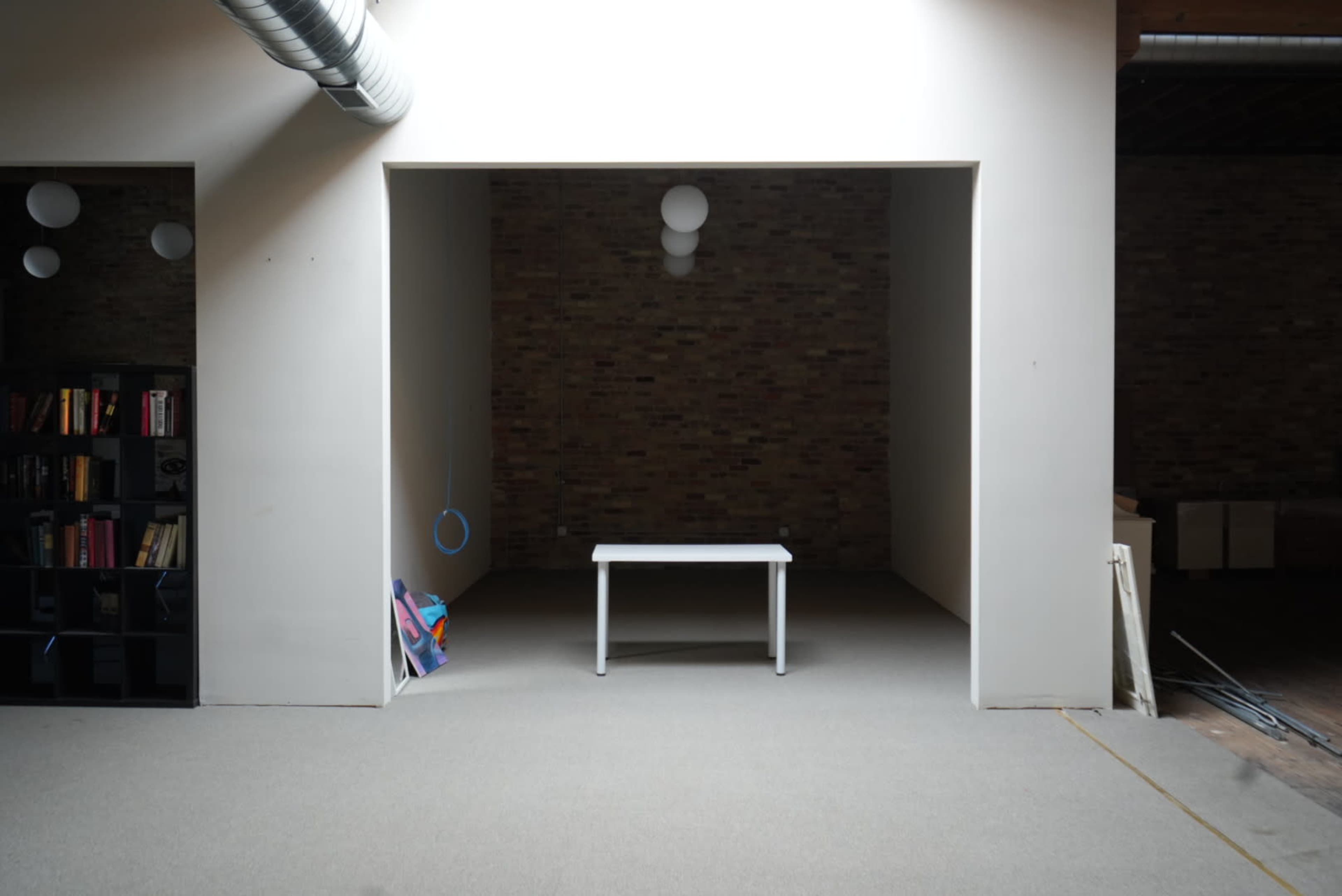 A small, sparsely furnished room features a white table centered on a carpeted floor, with brick walls and a bookshelf visible on one side.