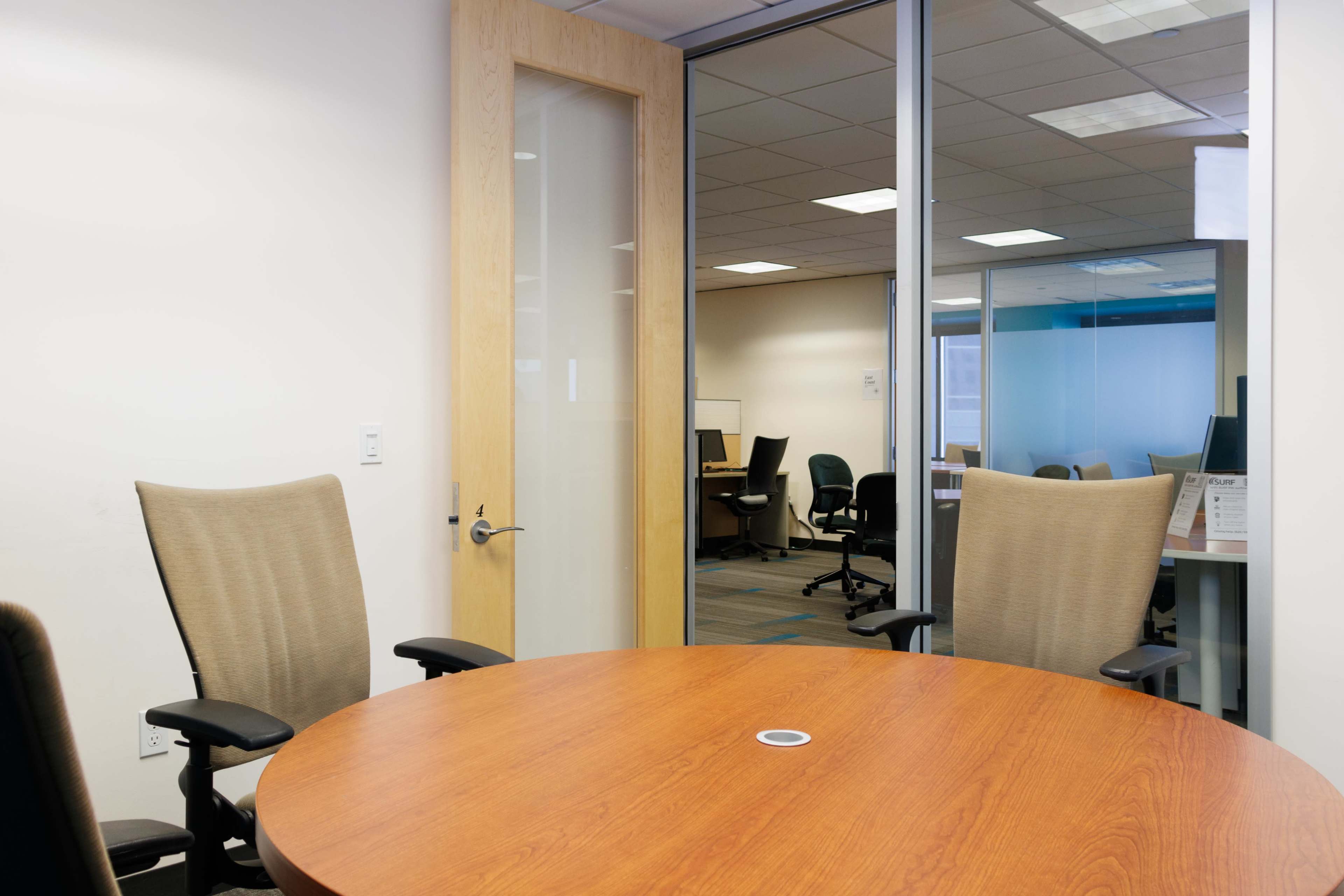 A conference room with a round wooden table and two empty chairs faces a glass wall that leads to an adjacent office area with desks and chairs.