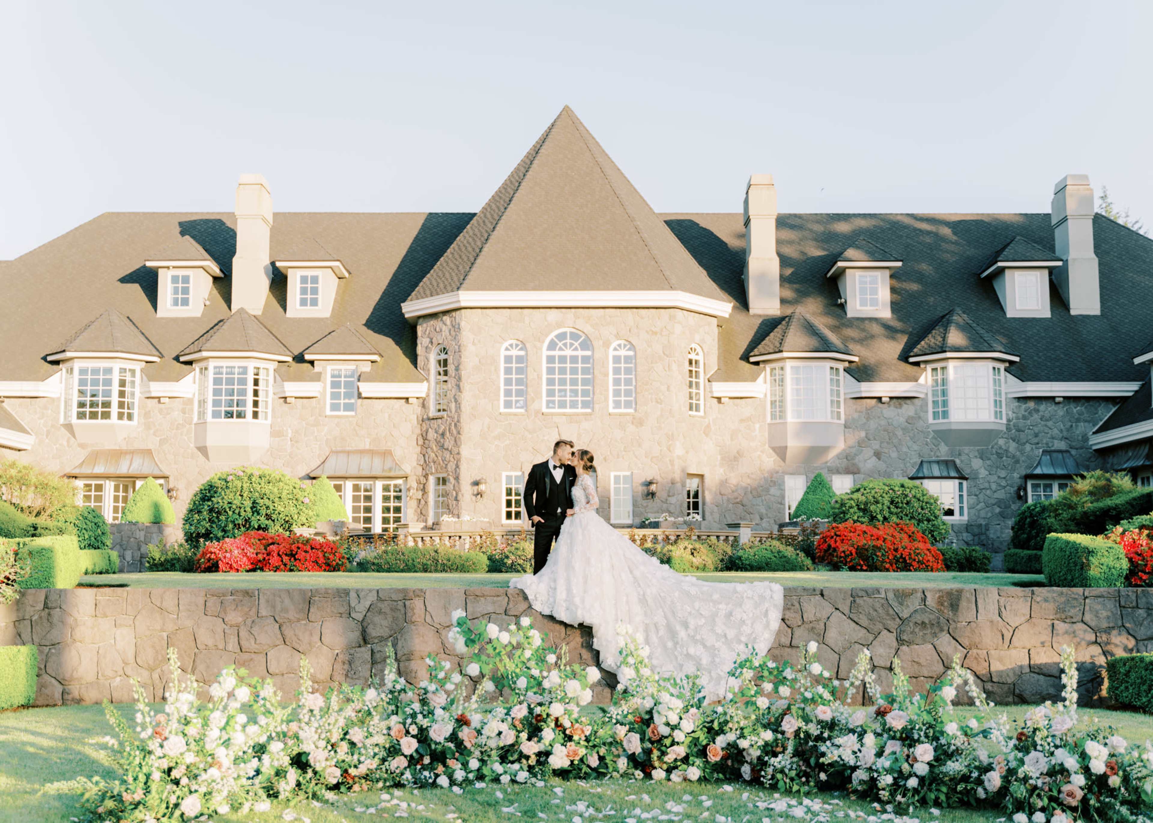 A couple in wedding attire stands together on a stone terrace in front of a large, intricately designed mansion surrounded by well-maintained flower beds.
