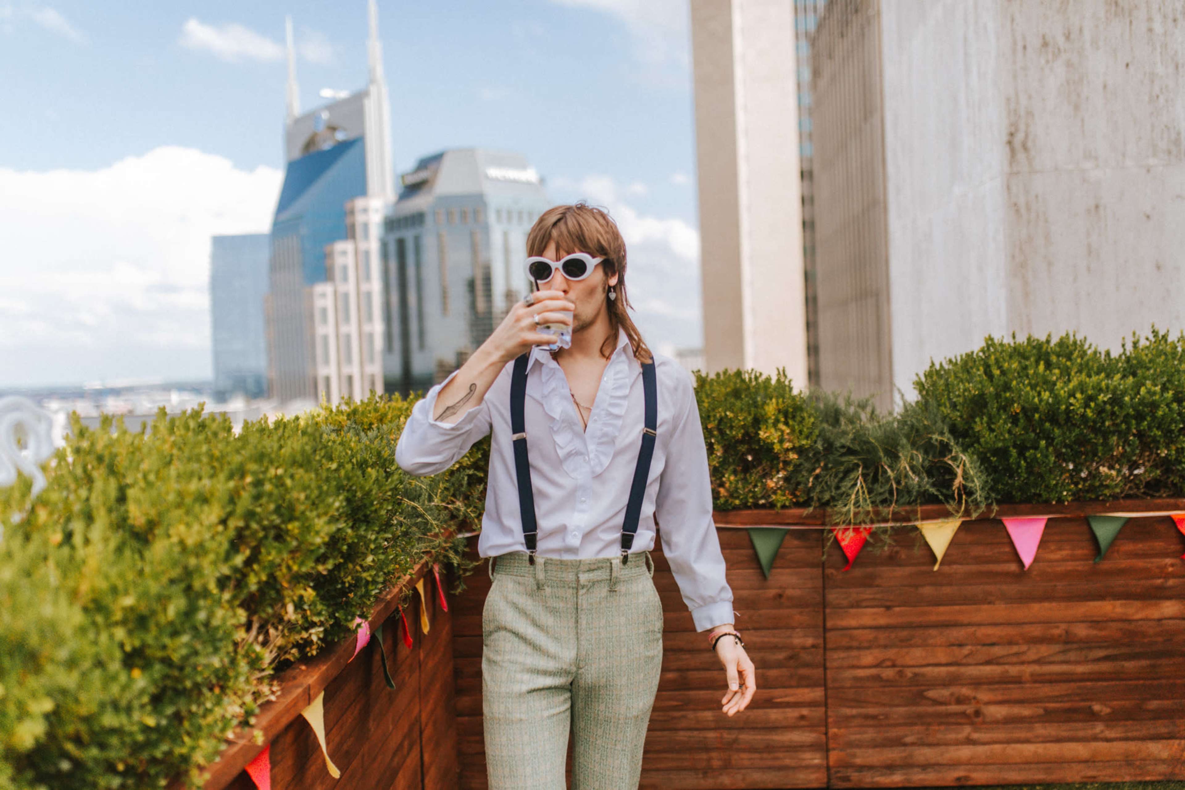 A person wearing sunglasses and a shirt with suspenders holds a drink while walking on a rooftop garden with city skyscrapers in the background.