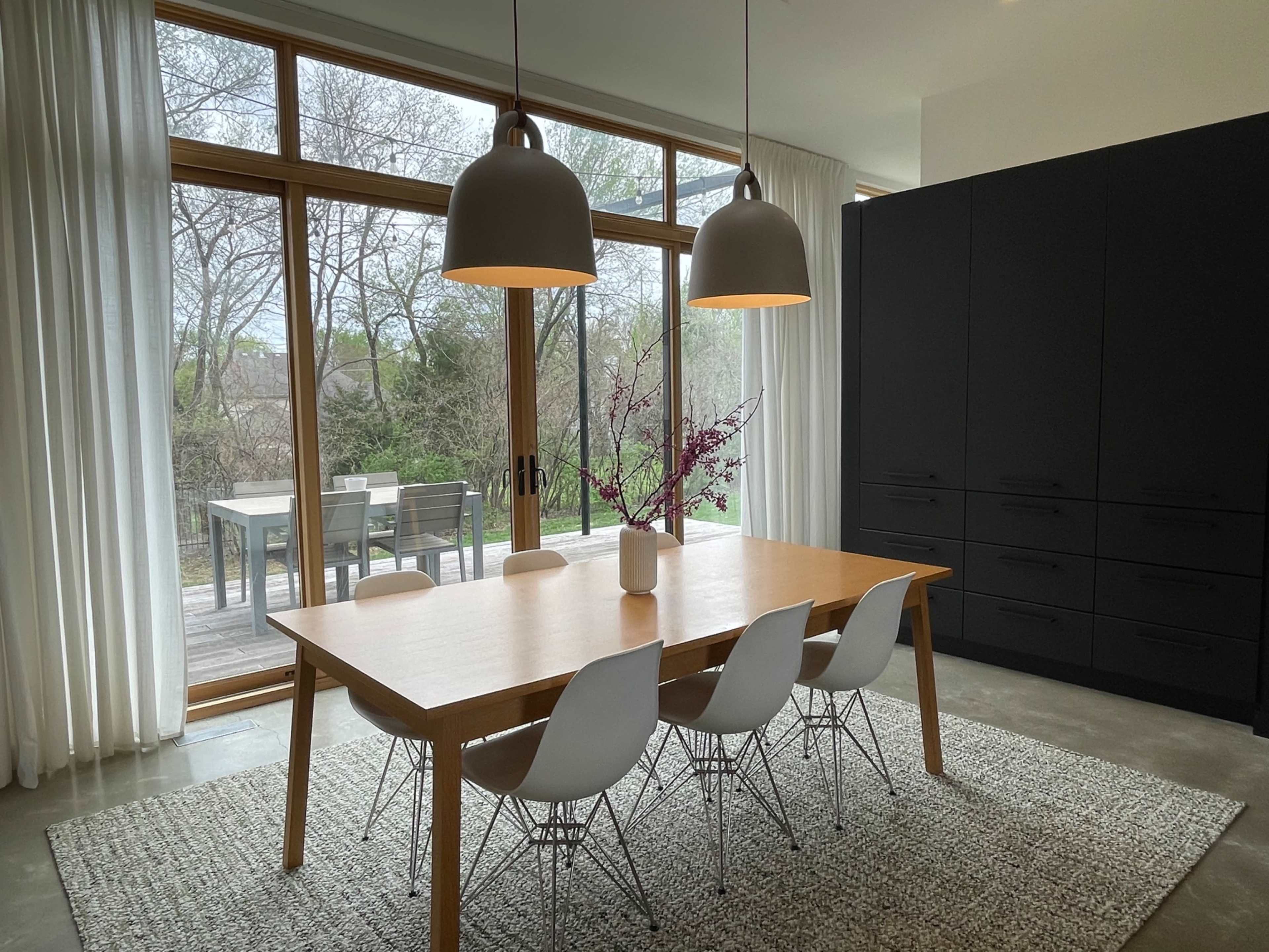 A modern dining area features a wooden table surrounded by white chairs, with large windows overlooking a green outdoor space.