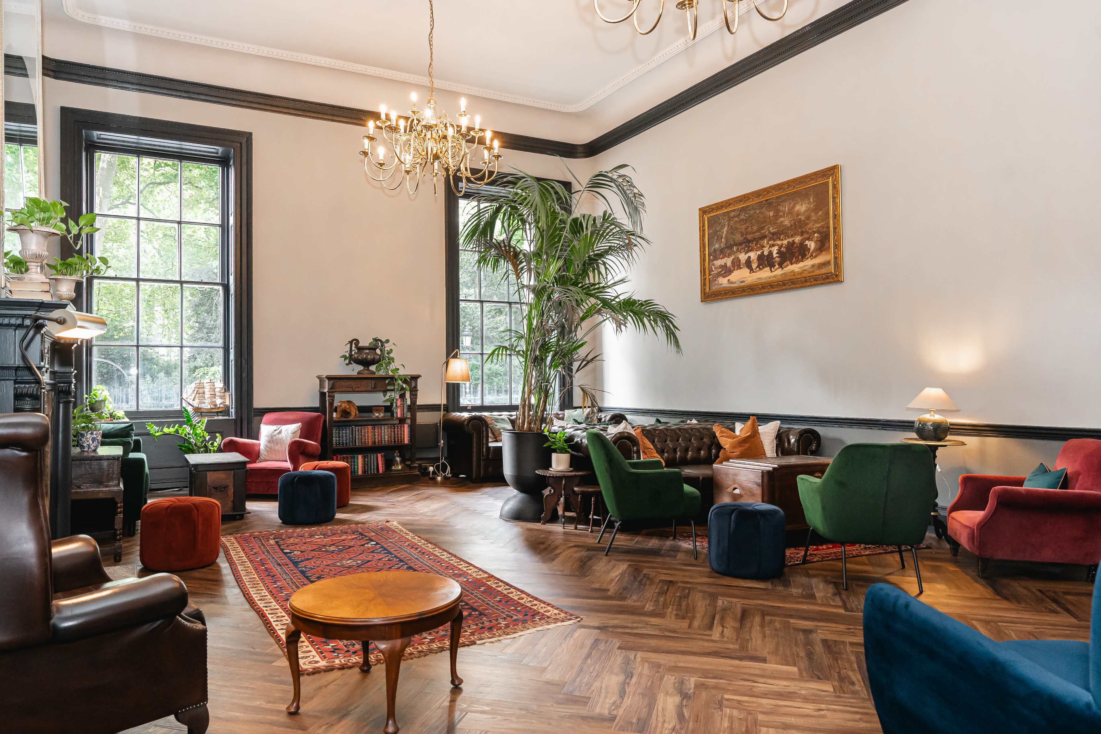The image depicts a cozy lounge area with a mix of upholstered chairs, a wooden table, and various plants, illuminated by natural light from large windows.